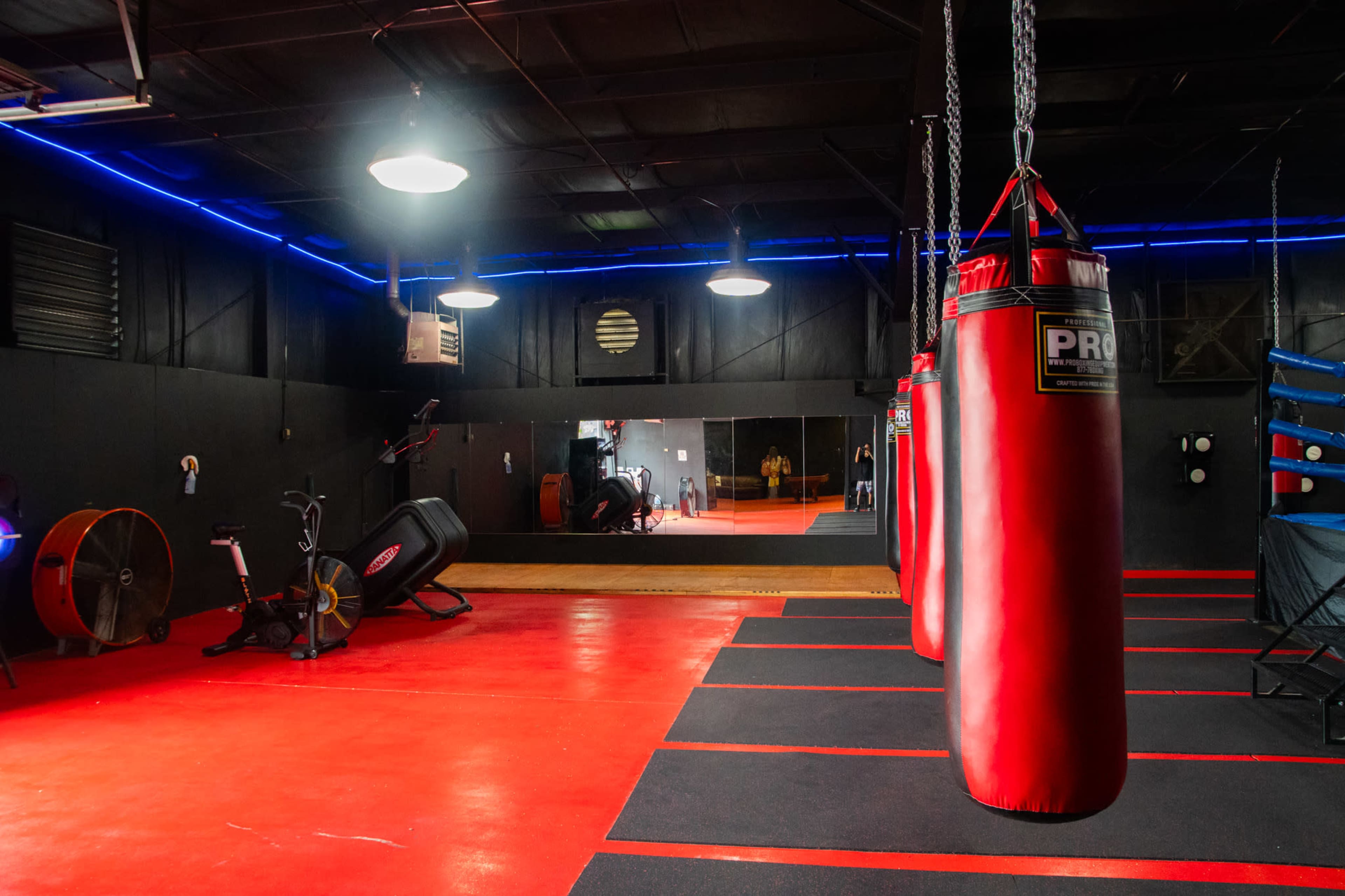 The image shows a boxing gym with red punching bags, red flooring, and a large mirror on the back wall.