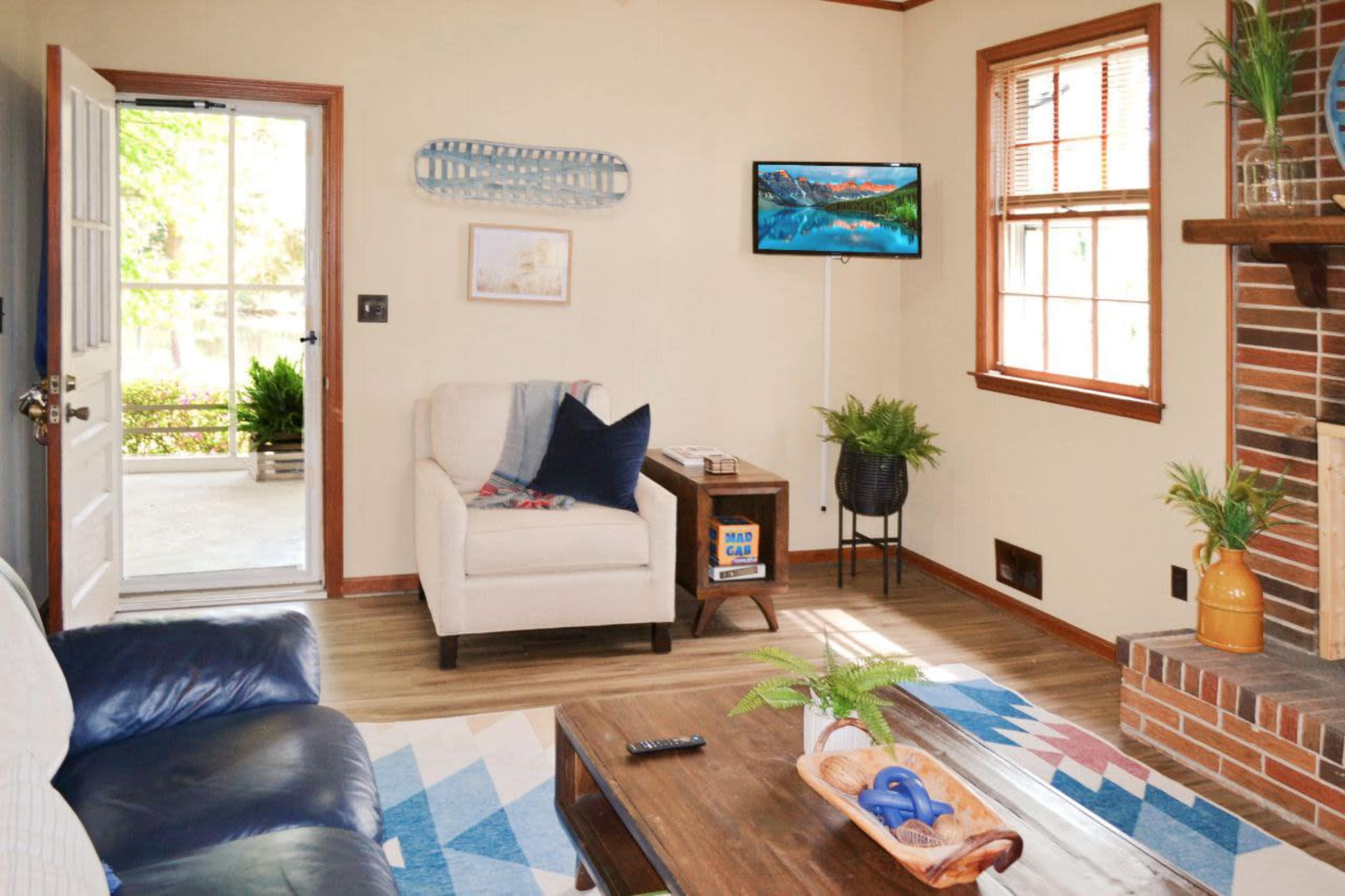 A cozy living room with a beige armchair, a blue couch, a wooden coffee table, a television mounted on the wall, and a view of a door leading outside.