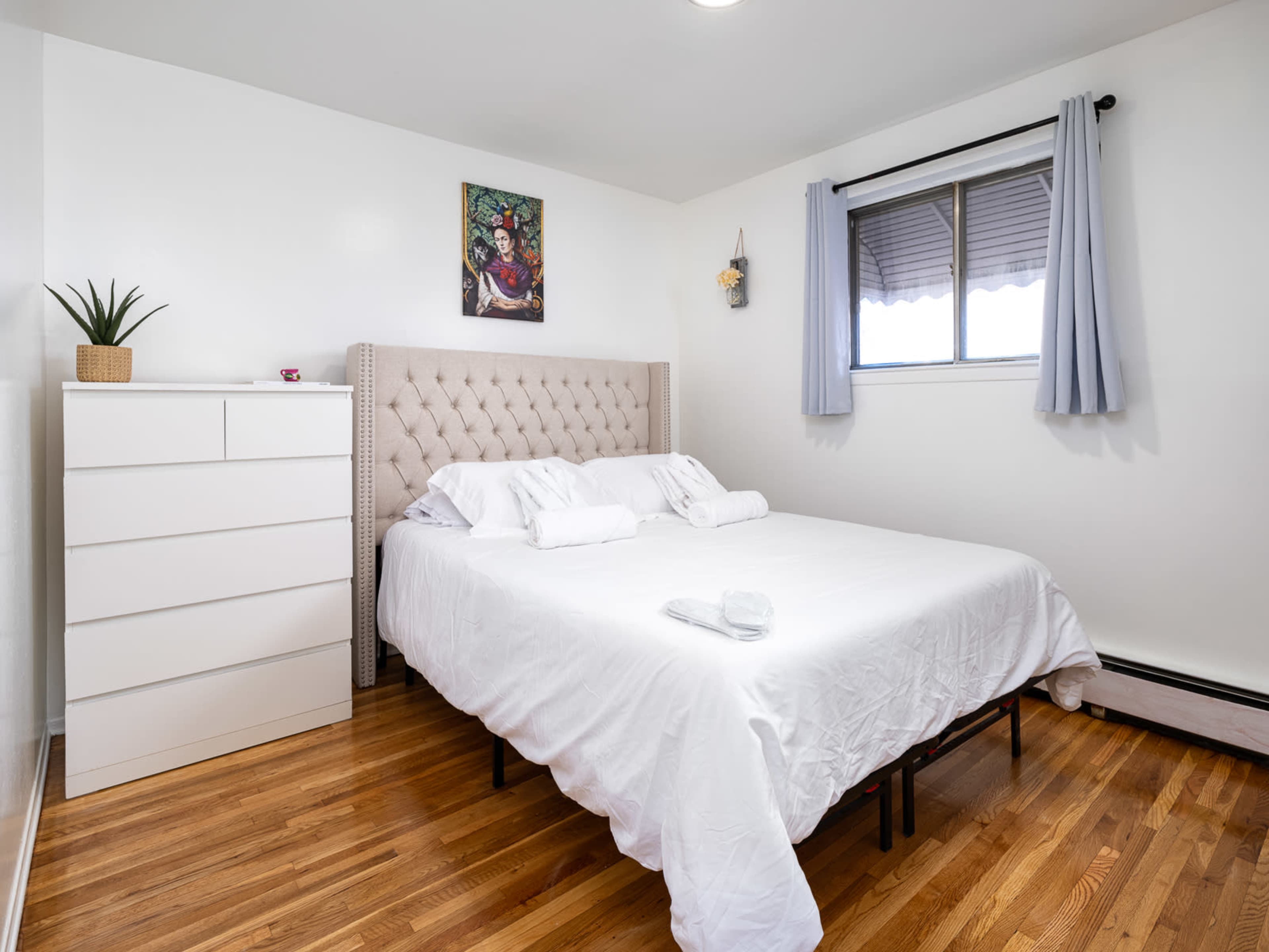 A neatly arranged bedroom features a queen-sized bed with a tufted headboard, a white dresser, and a window with light curtains.