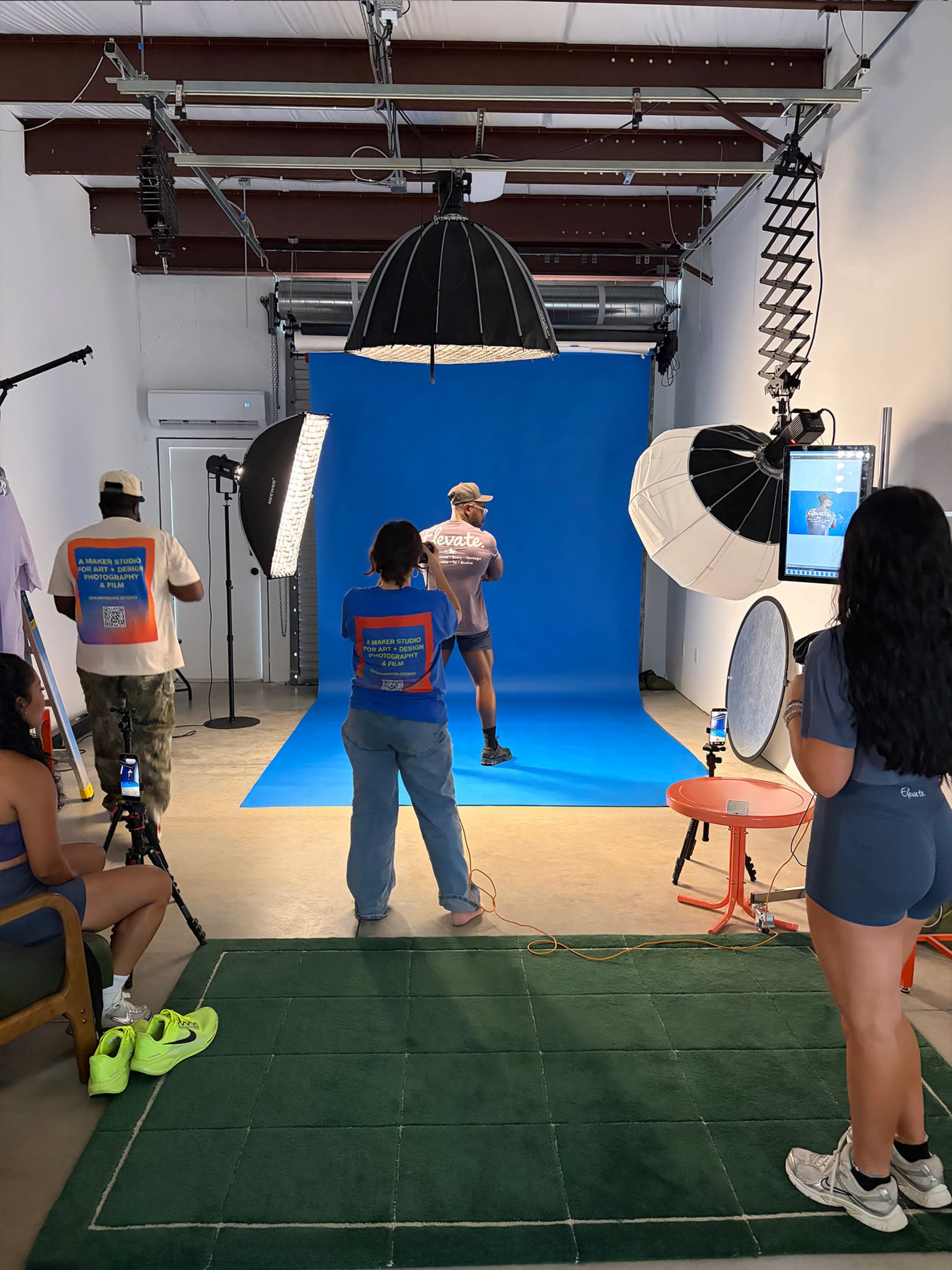 A photography studio features a model posing against a bright blue backdrop, surrounded by photographers and lighting equipment.