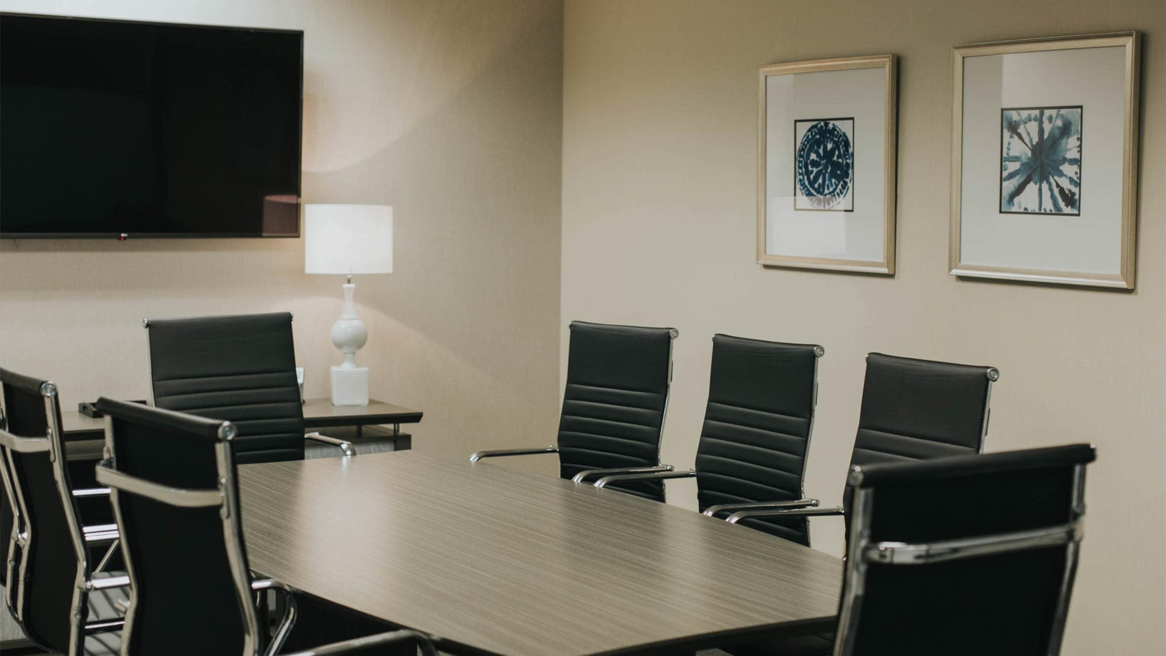A conference room features a large wooden table surrounded by six black chairs, a lamp, a TV mounted on the wall, and two framed artworks.