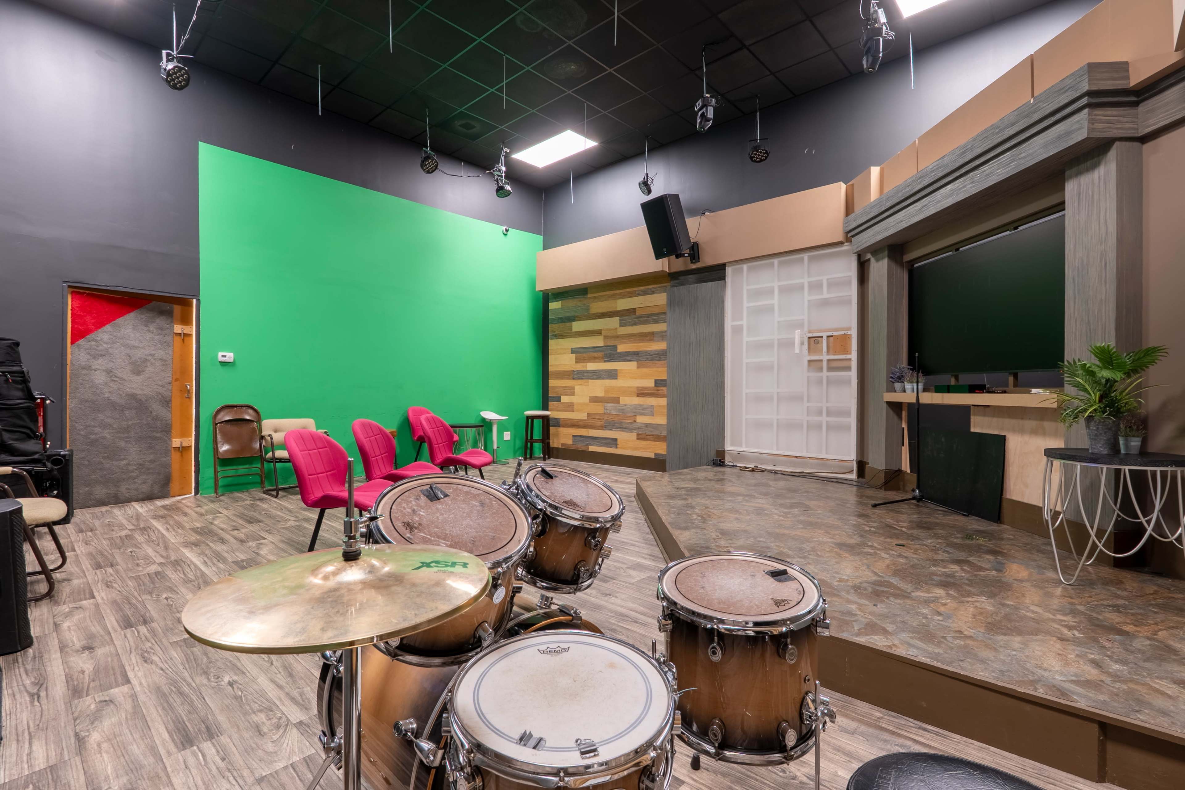 The image shows a rehearsal space featuring a drum set in the foreground, a green screen wall, and various seating arrangements in a modern studio setting.