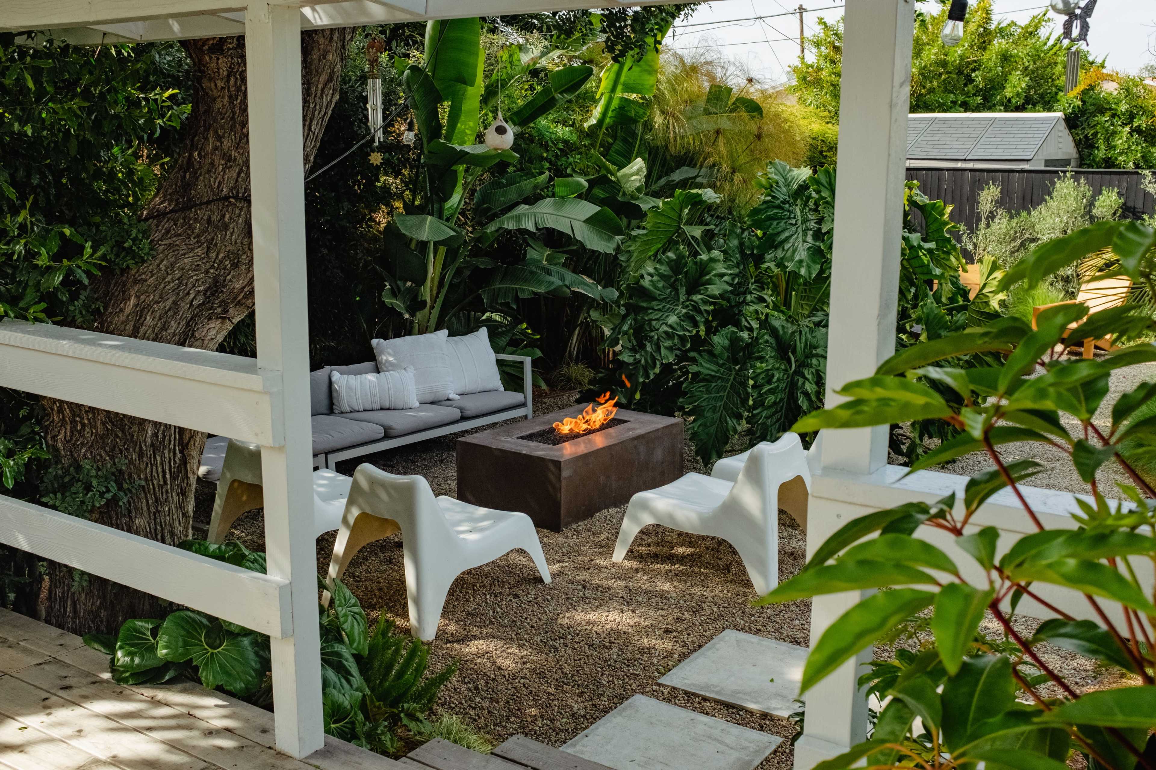 A cozy outdoor seating area features a fire pit surrounded by four white chairs and lush greenery.
