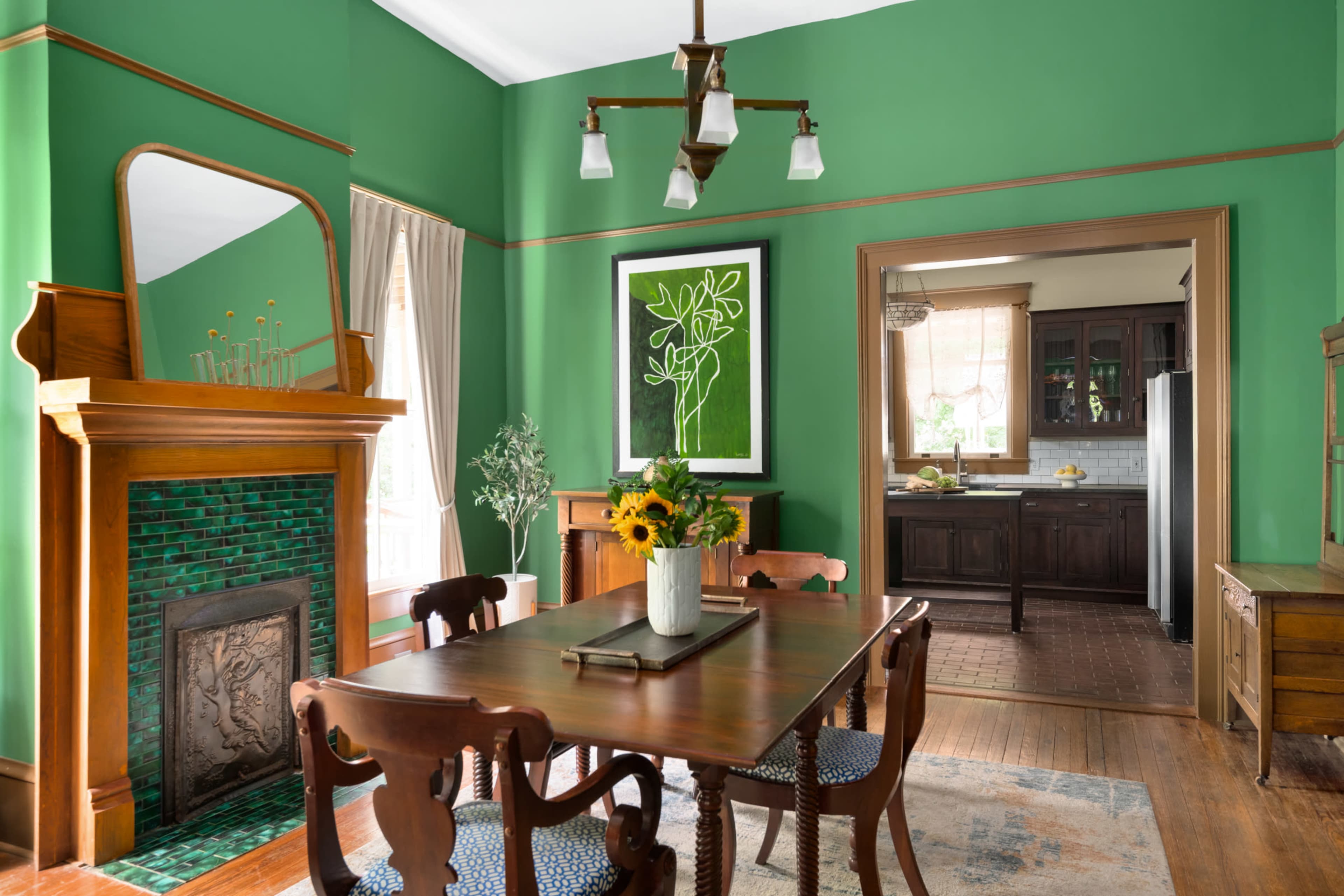A dining room with a green wall, wooden dining table, and a fireplace features a mirror above it and leads to a kitchen area visible through an open doorway.
