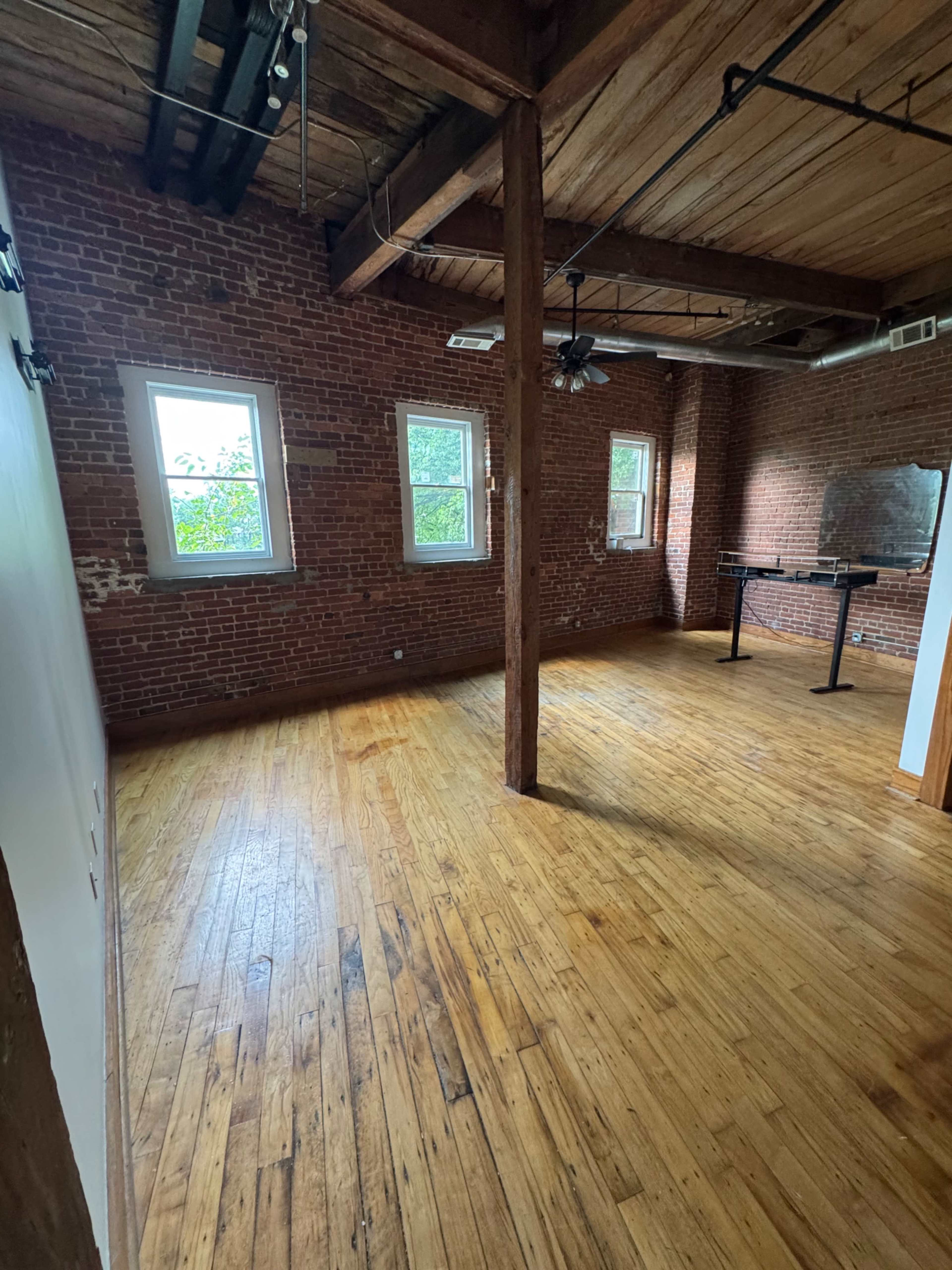 The image shows a spacious room with wooden flooring, exposed brick walls, and large windows, featuring a ceiling fan and a mirrored wall on one side.