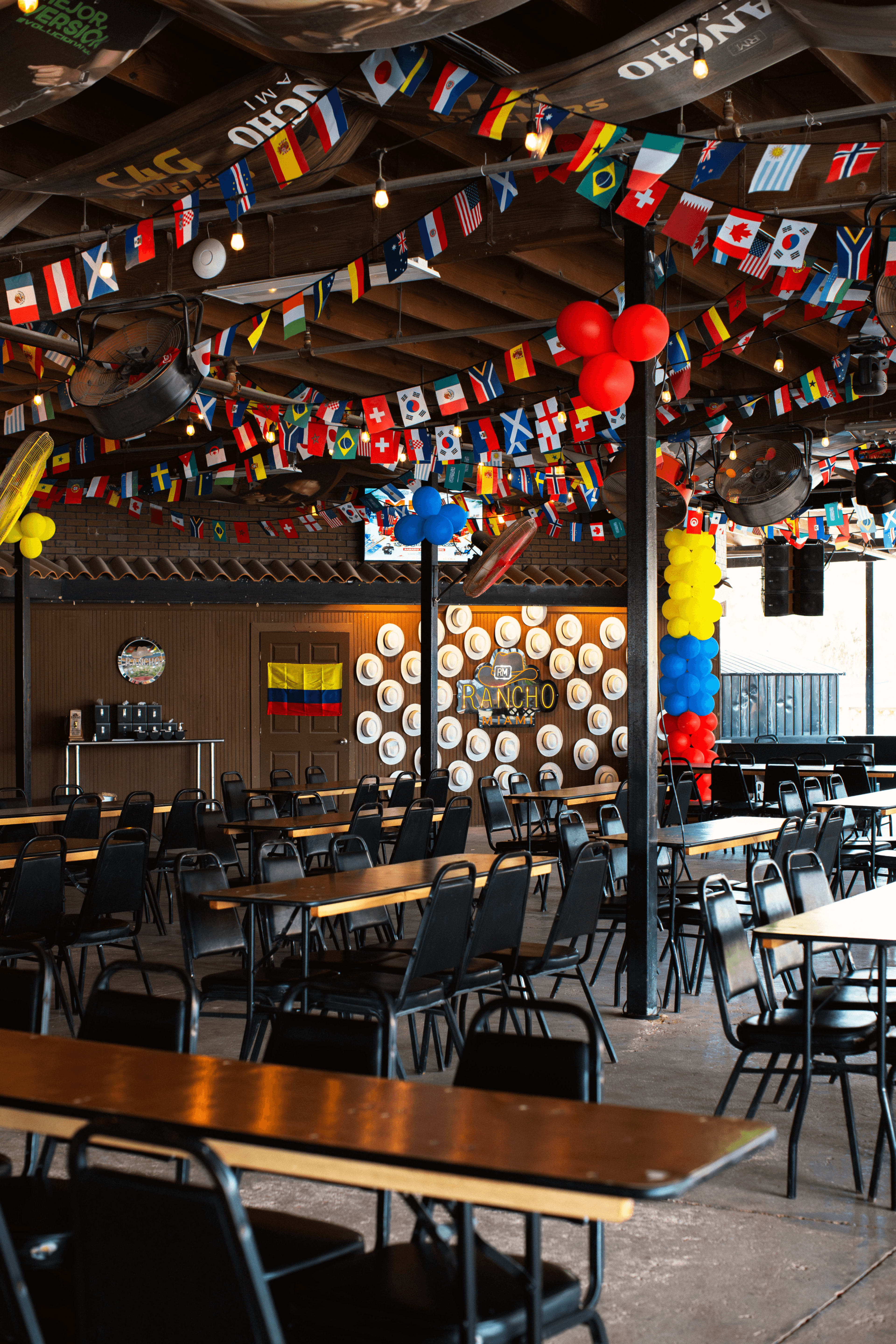 The interior of a restaurant features long tables and chairs arranged in rows beneath a ceiling adorned with colorful flags and balloons.