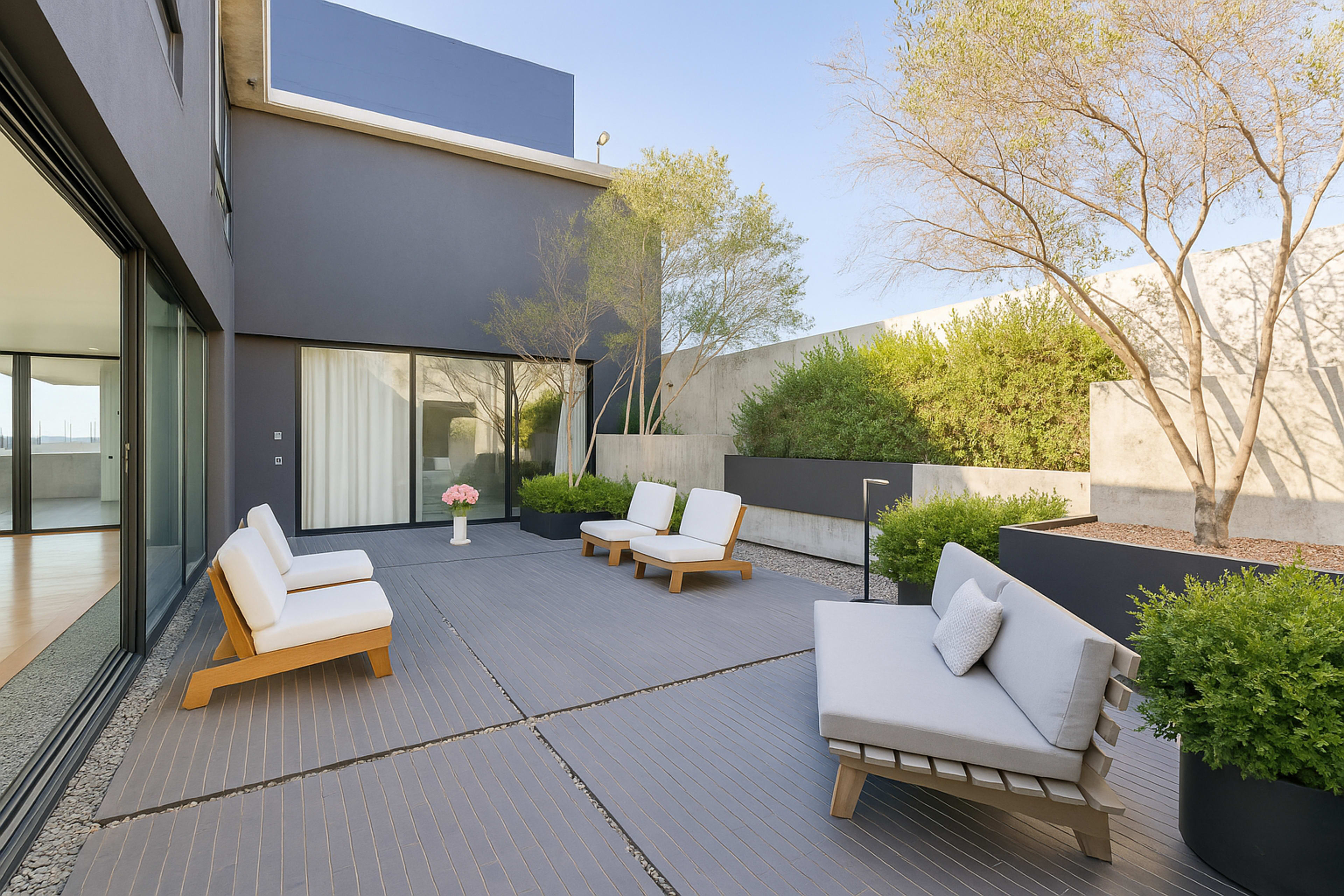 A modern patio features minimalist seating, planters, and textured flooring surrounded by greenery and a gray wall.