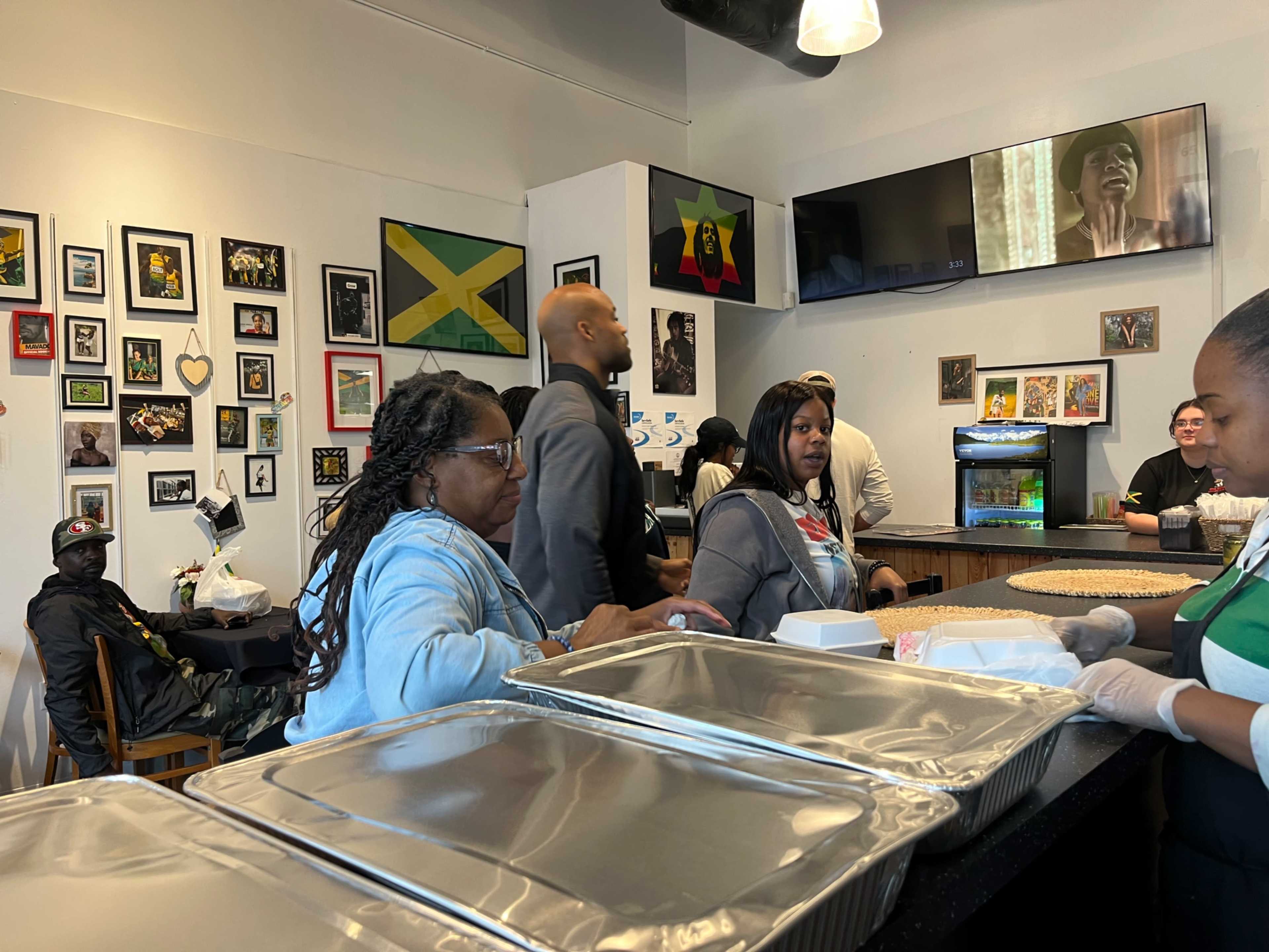 A busy restaurant scene features patrons enjoying their meals while staff at the counter serve food in front of a wall adorned with pictures and flags.