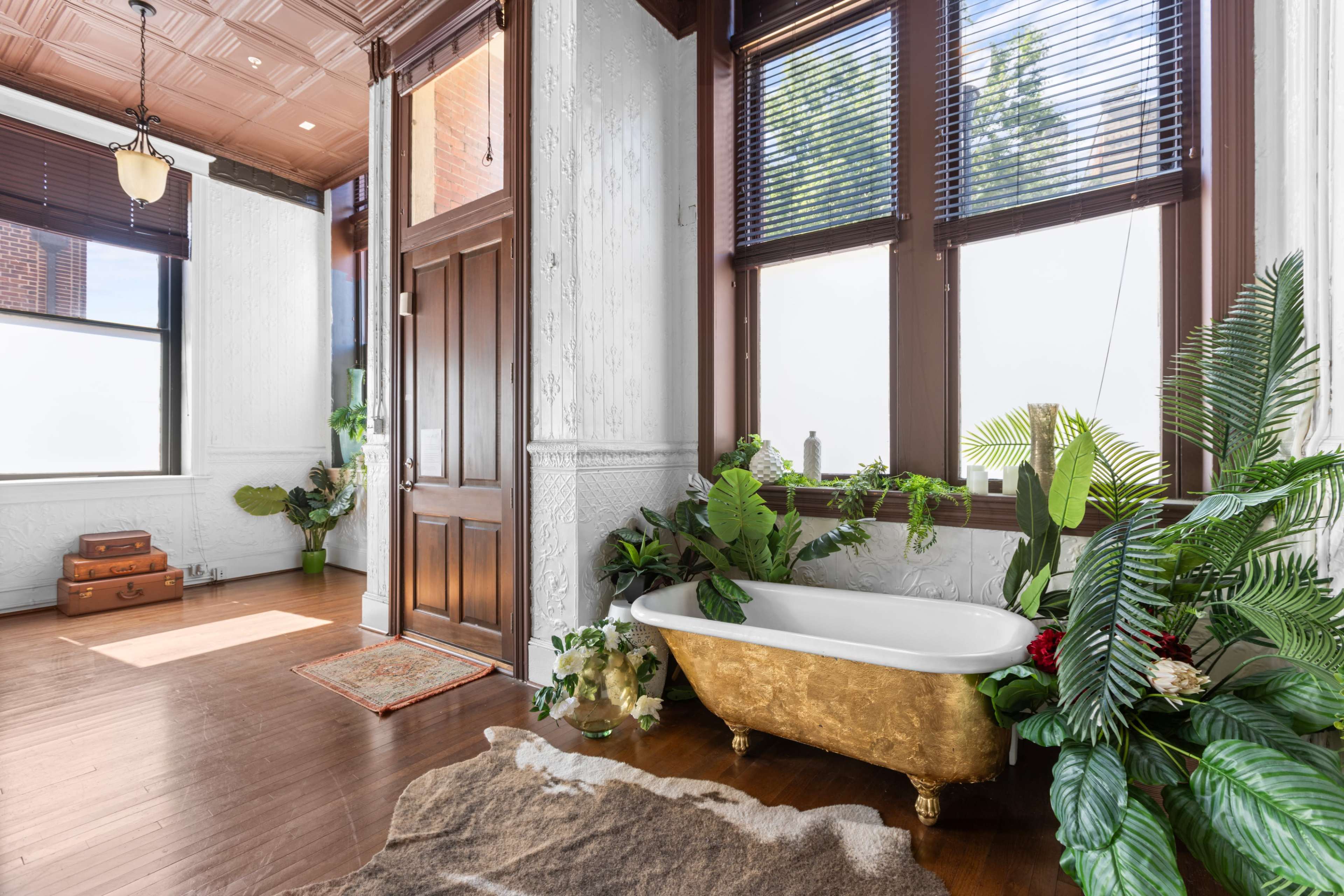 A vintage gold clawfoot bathtub is positioned near large windows, surrounded by various indoor plants in a sunlit room with wooden flooring and decorative moldings.