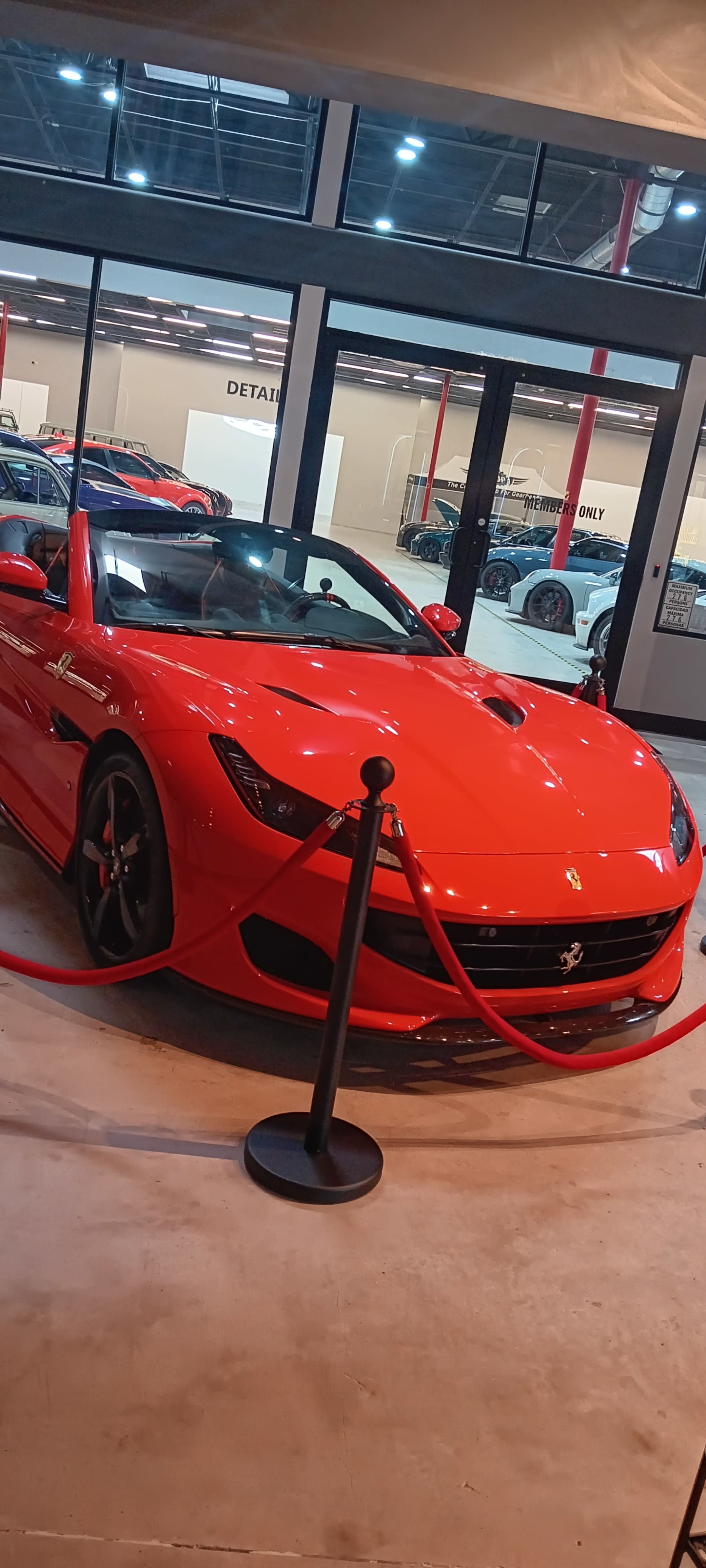 A bright red convertible sports car is displayed behind a velvet rope in a showroom space.
