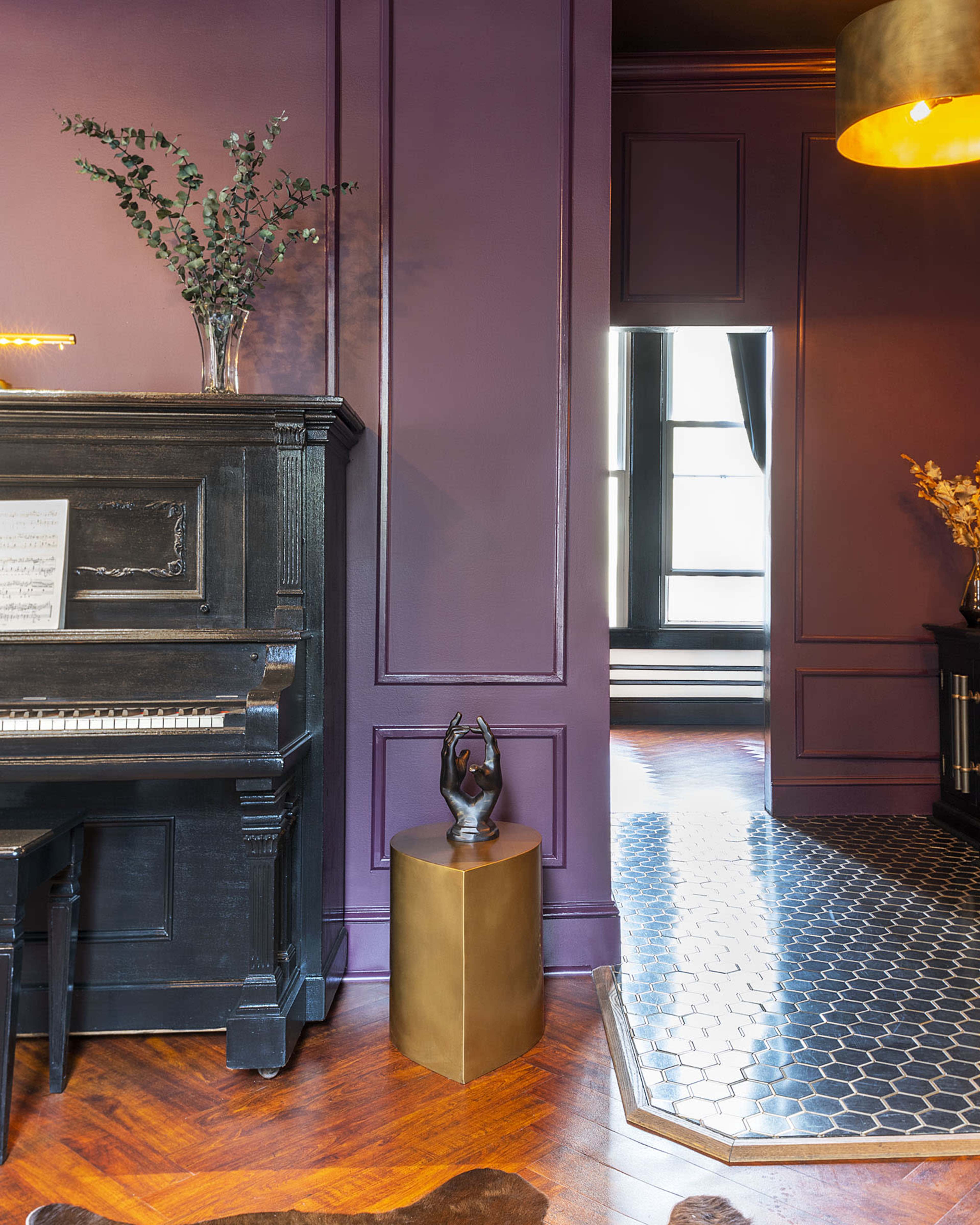 A dark-colored piano positioned against a purple wall, with a small brass table beside it and a doorway leading to another room in the background.