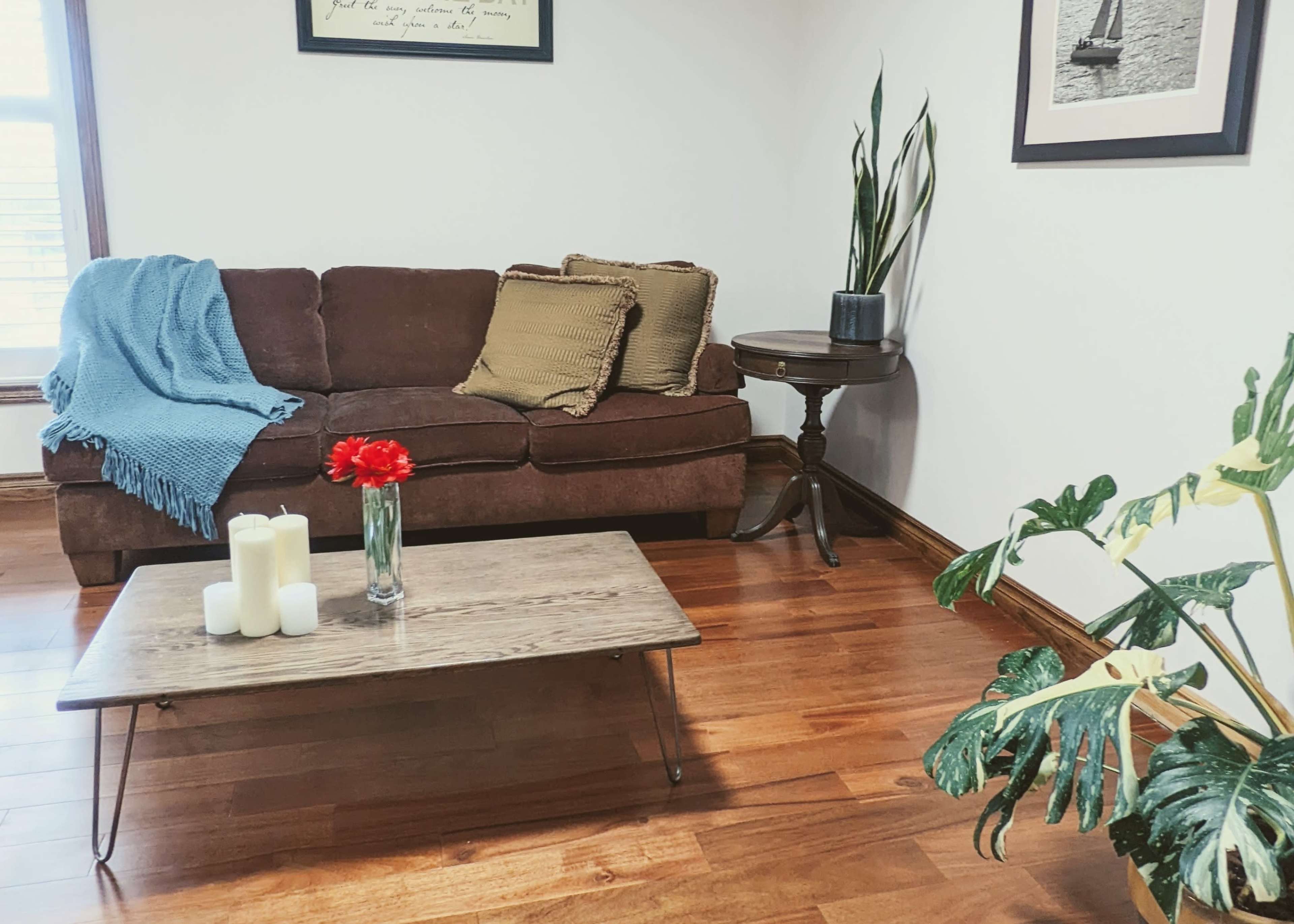 A brown couch with throw pillows sits near a wooden coffee table topped with candles and a flower vase, all placed on a hardwood floor in a well-lit room.