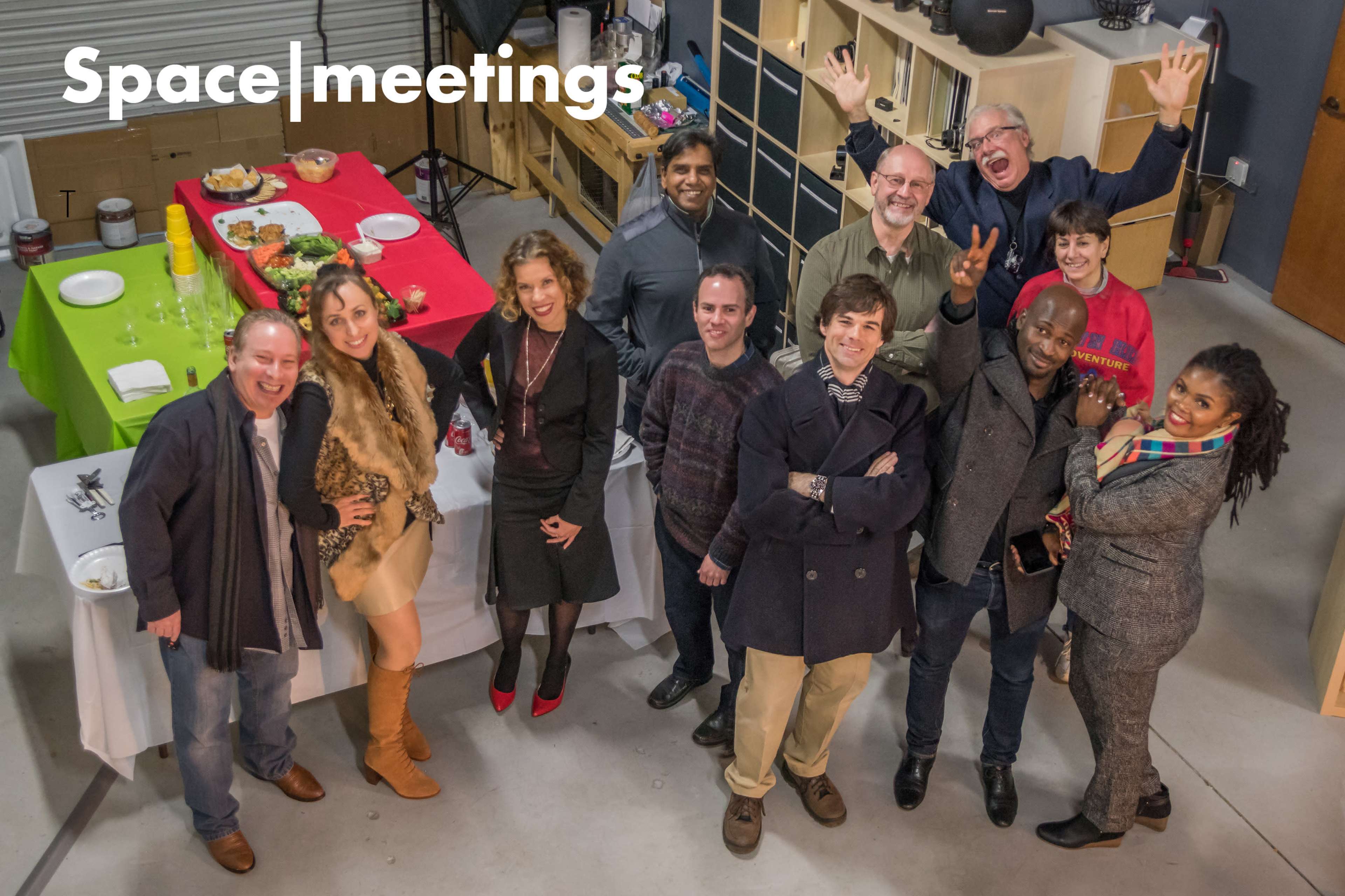 A group of twelve people is gathered in a warehouse, smiling and posing for the camera with food and drinks on a table in the background.