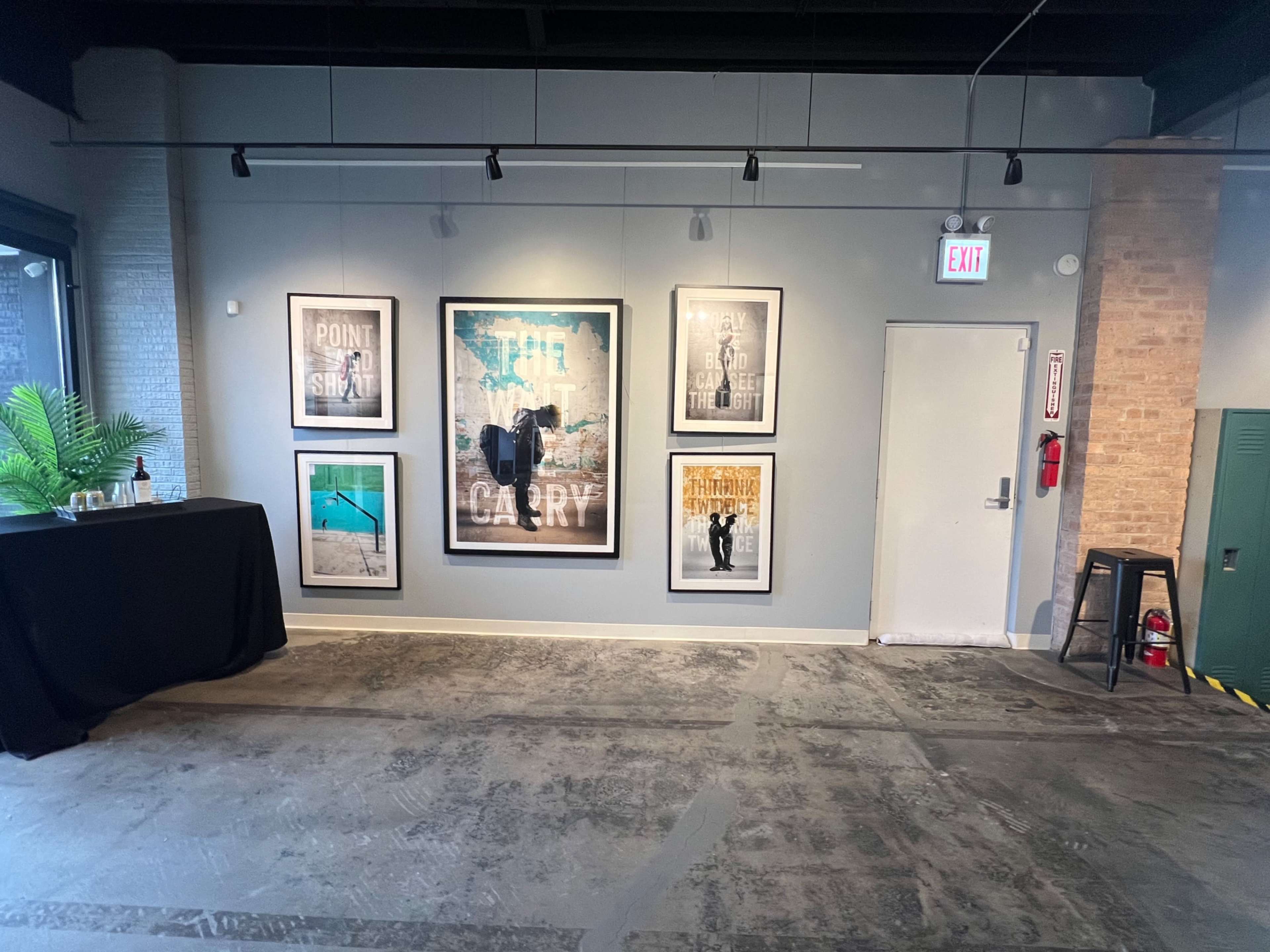 The image shows a wall in a gallery with six framed posters and a black table in the corner.
