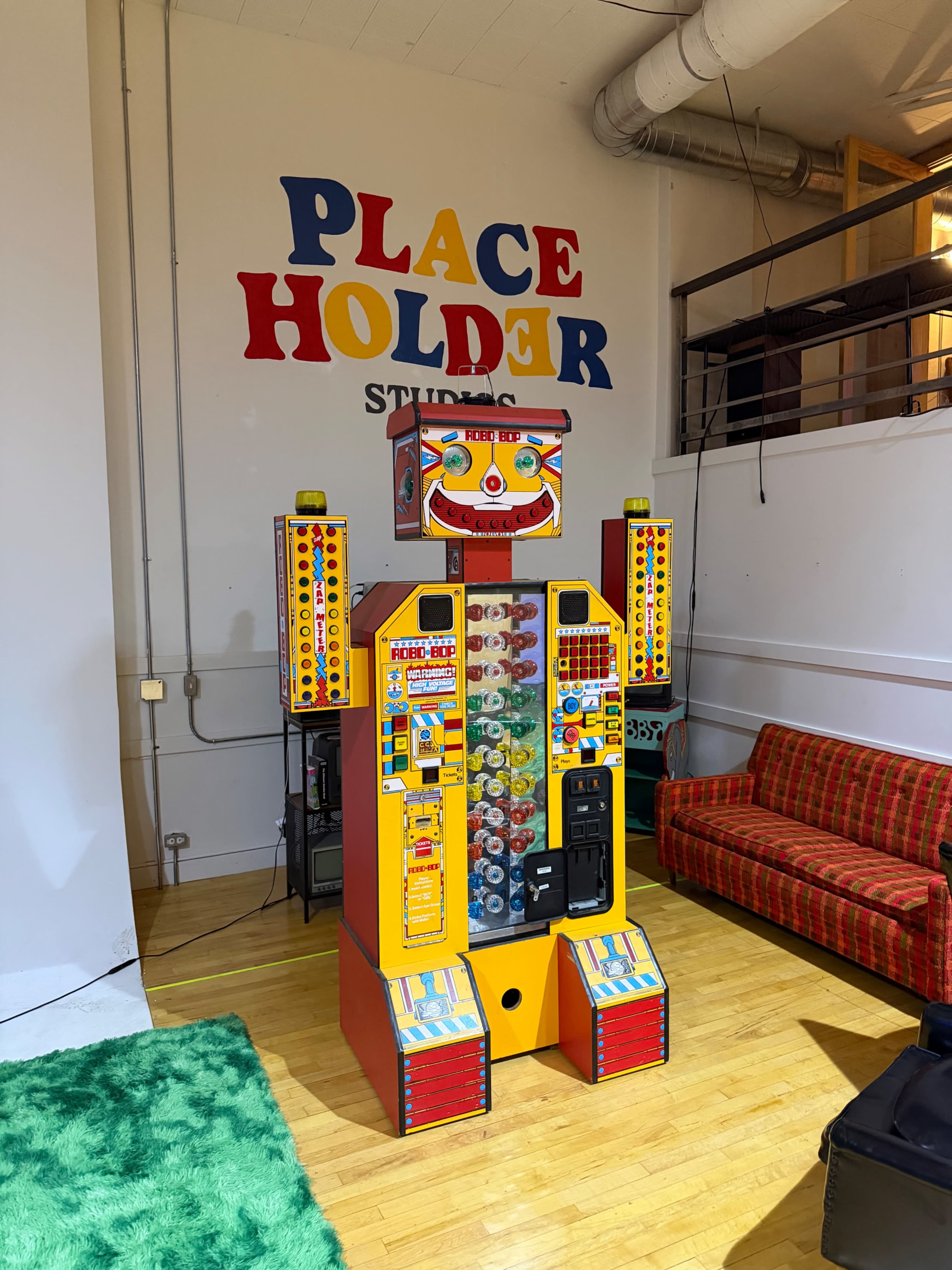 The image shows a colorful vintage-style vending machine robot standing in an open studio space with a bright green rug and a red couch in the background.