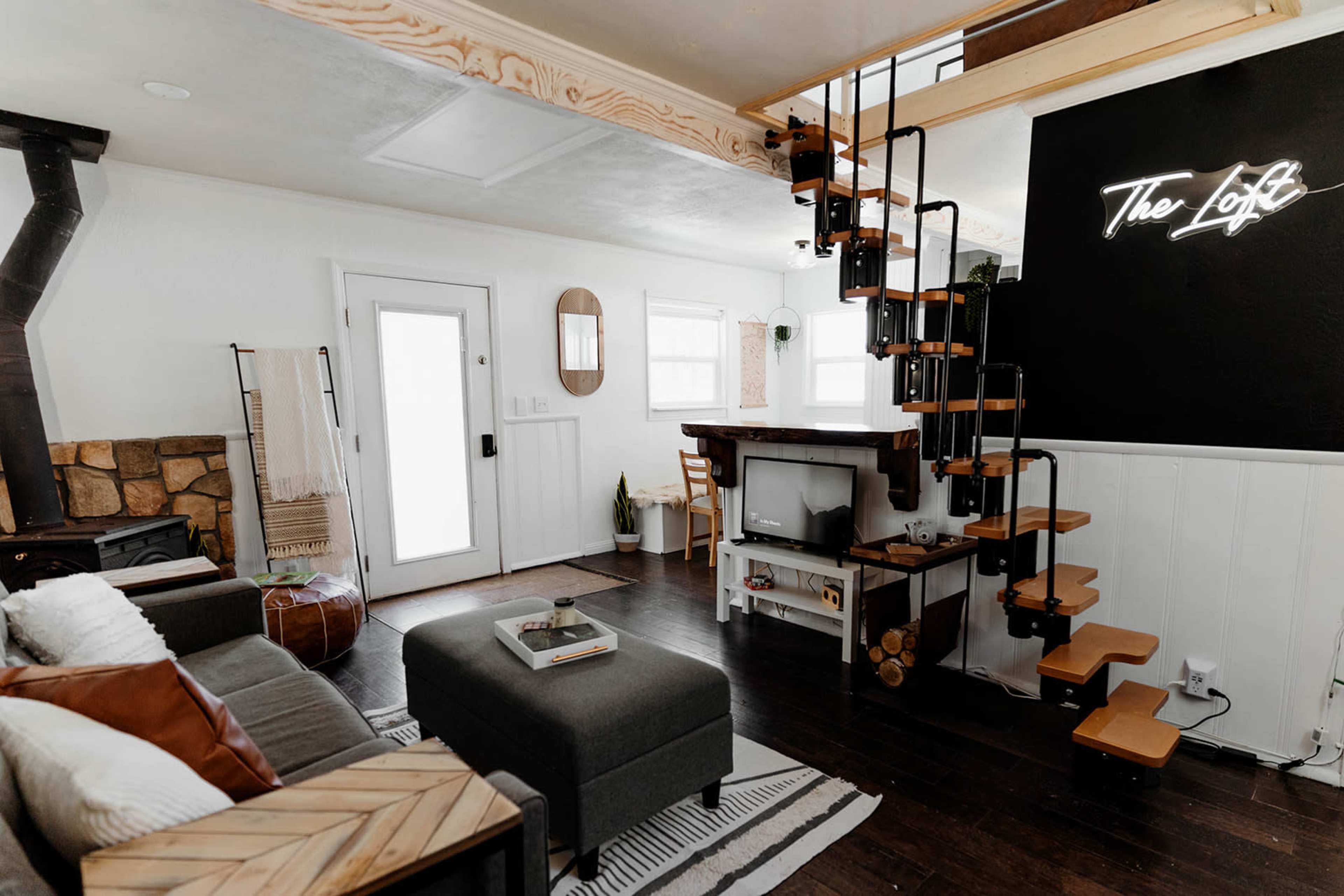 A modern living space featuring a cozy seating area, a staircase leading to a loft, and a black wall with a neon sign that reads "The Loft."