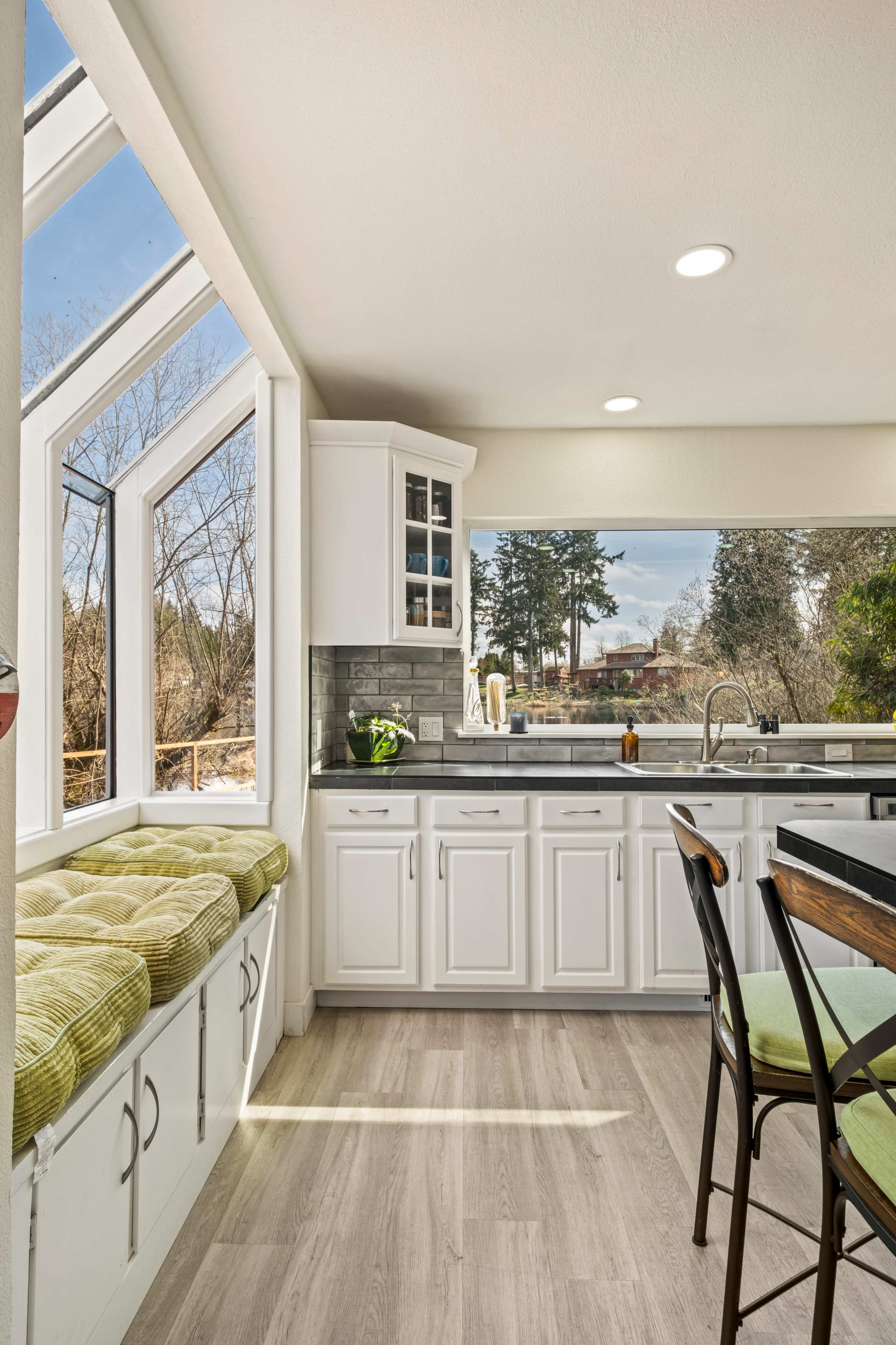 The image shows a modern kitchen featuring white cabinets, a sink area with a large window, and a green cushioned bench near the window.