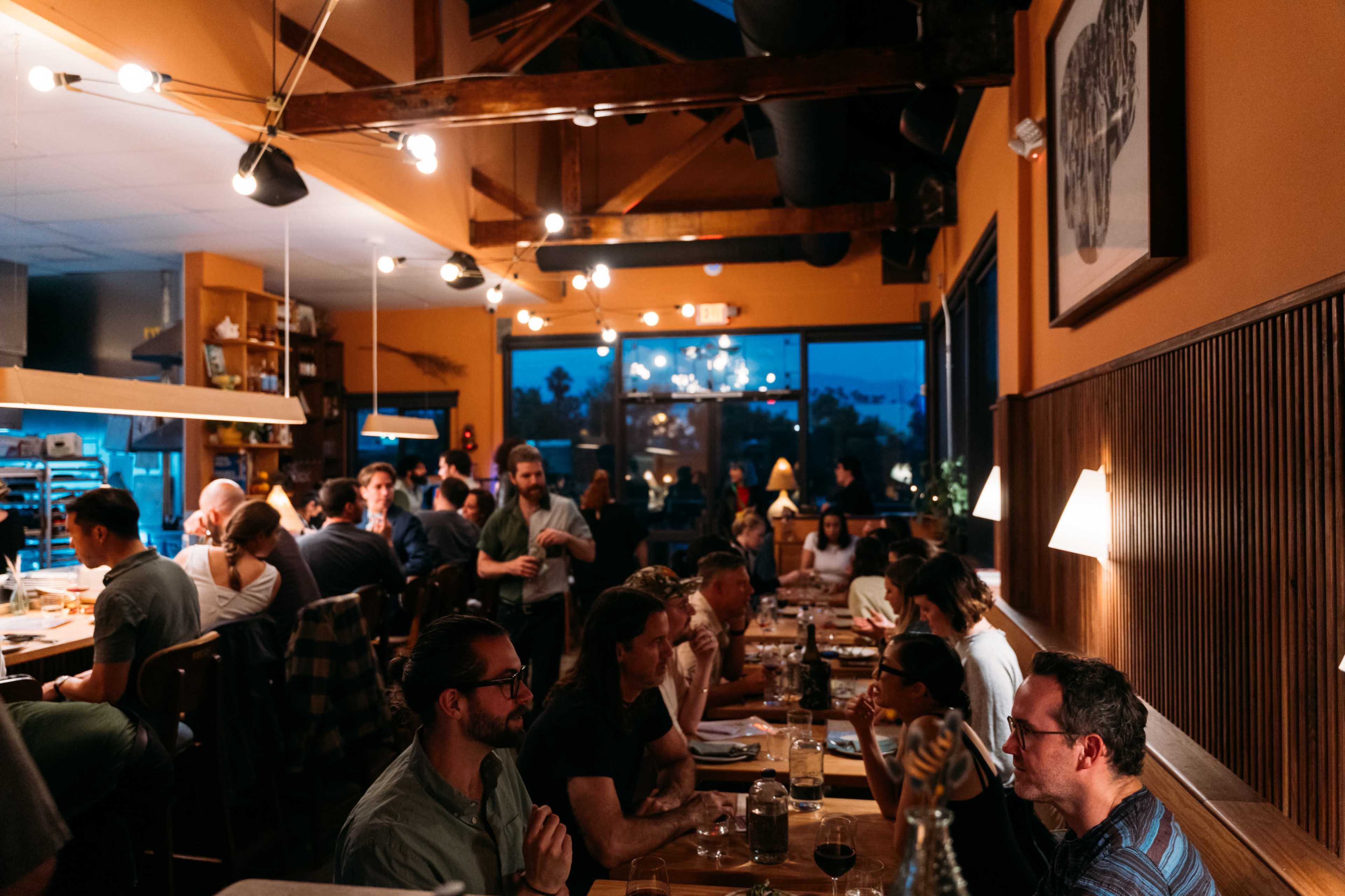 The image shows a busy restaurant interior with diners engaged in conversation, illuminated by warm lights and window views of the evening sky.