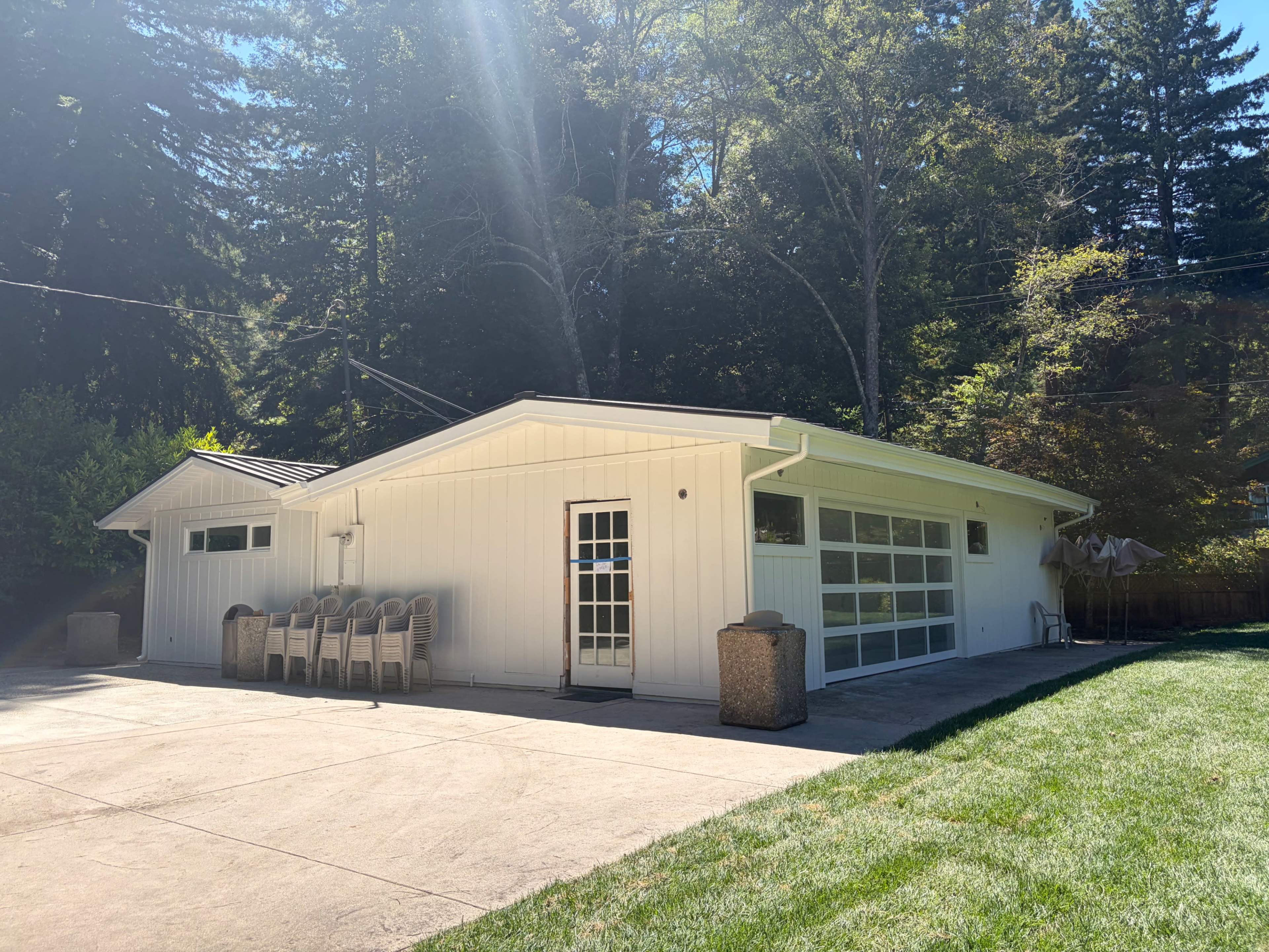 The image shows a modern white building with a large glass door and a patio area featuring six stacked chairs, surrounded by trees.