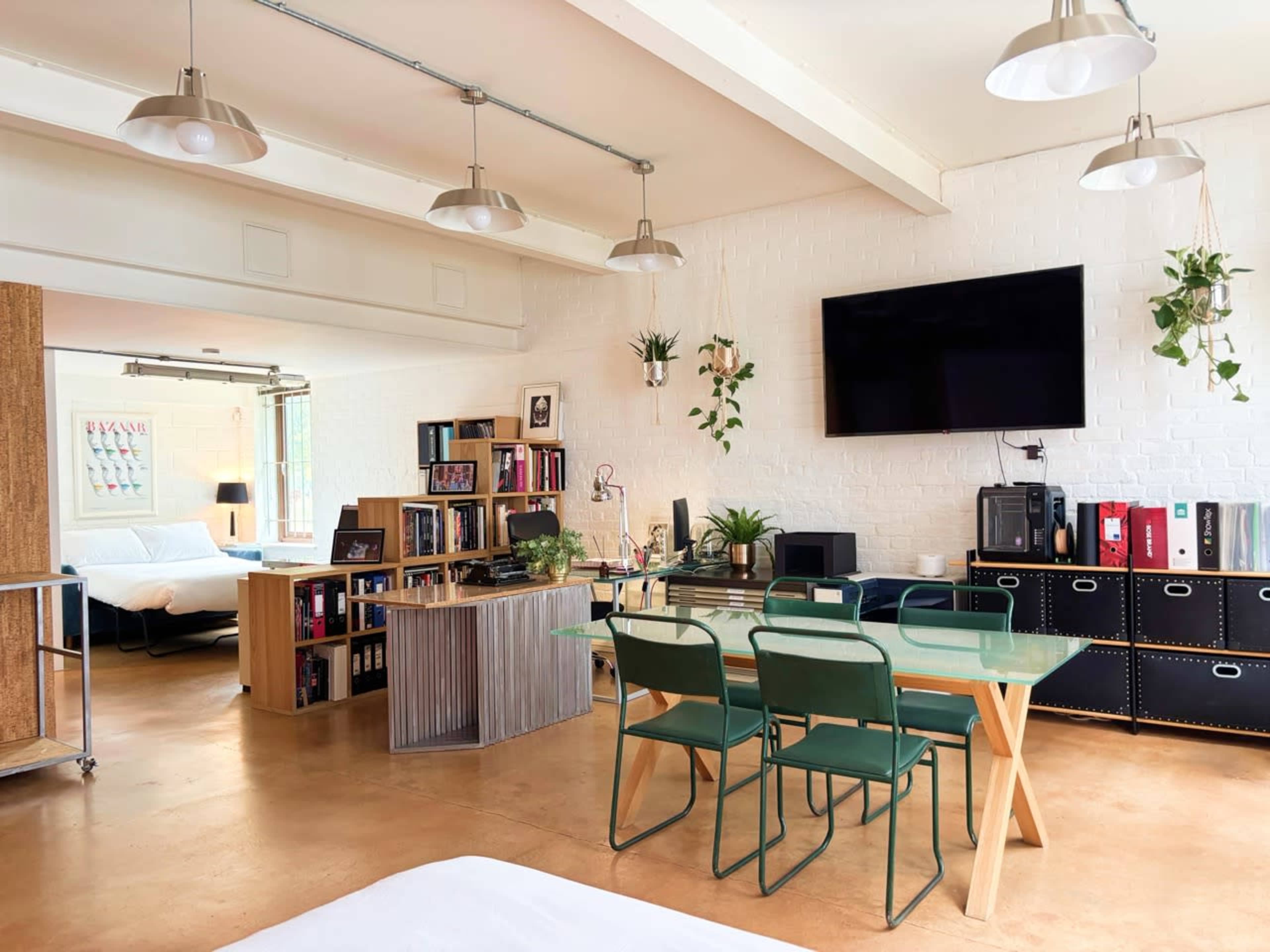 The image shows a contemporary living space with a dining area featuring a glass table and green chairs, a bookshelf, and a television mounted on a white brick wall.