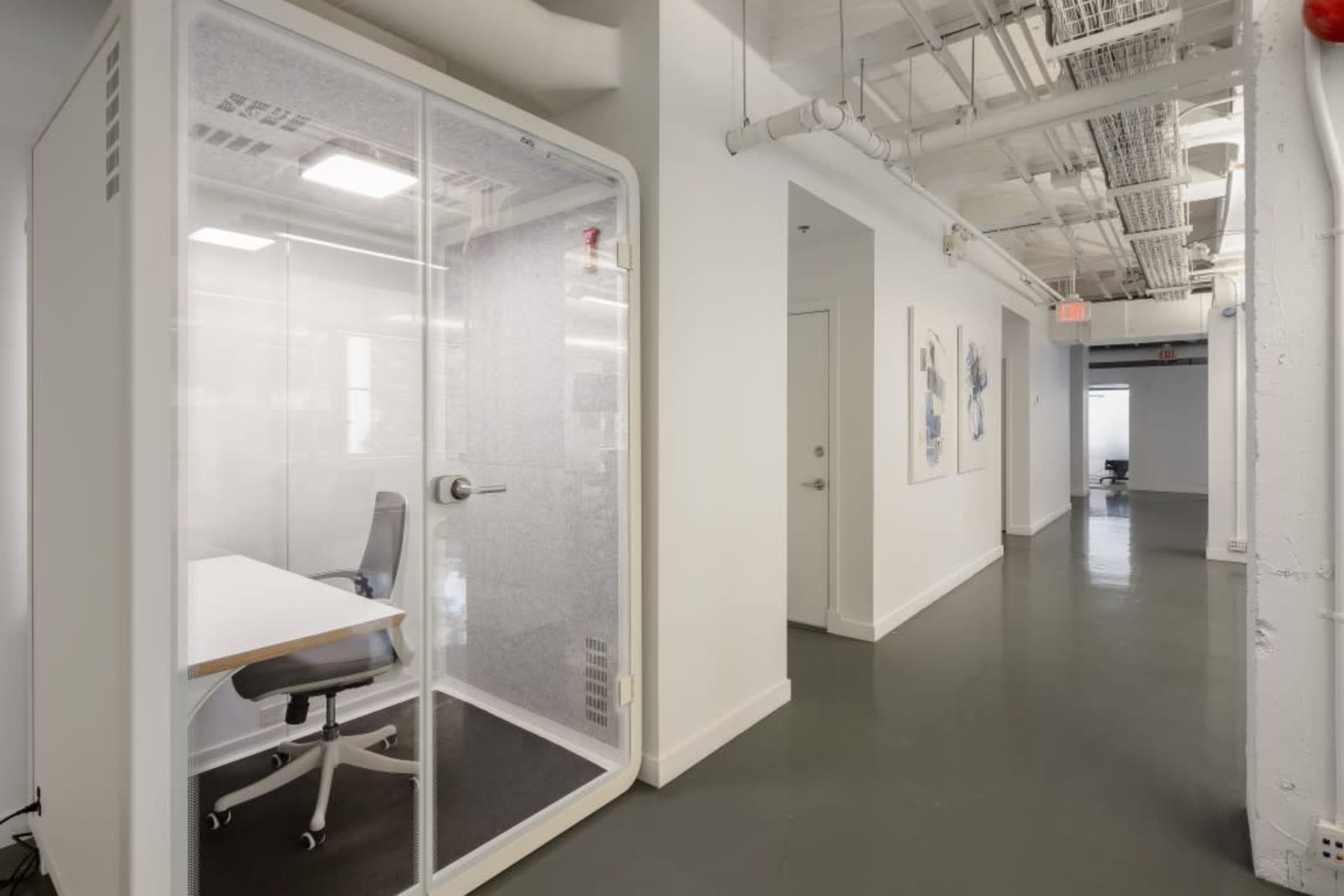 The image shows a modern office corridor with a glass-enclosed meeting room on one side and artwork on the walls.