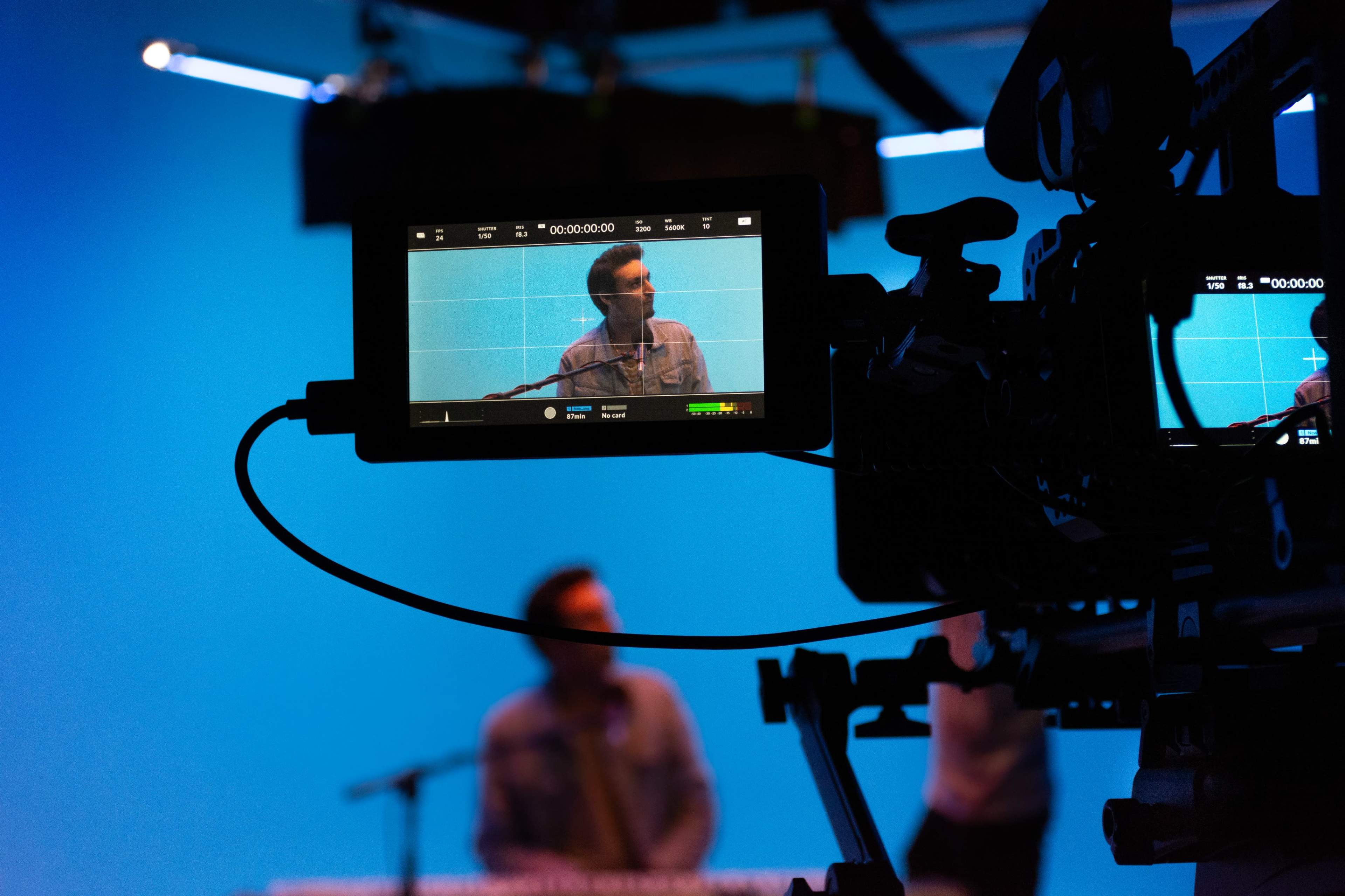 A camera displays a recording of a person speaking against a blue backdrop while another individual is visible in the background.