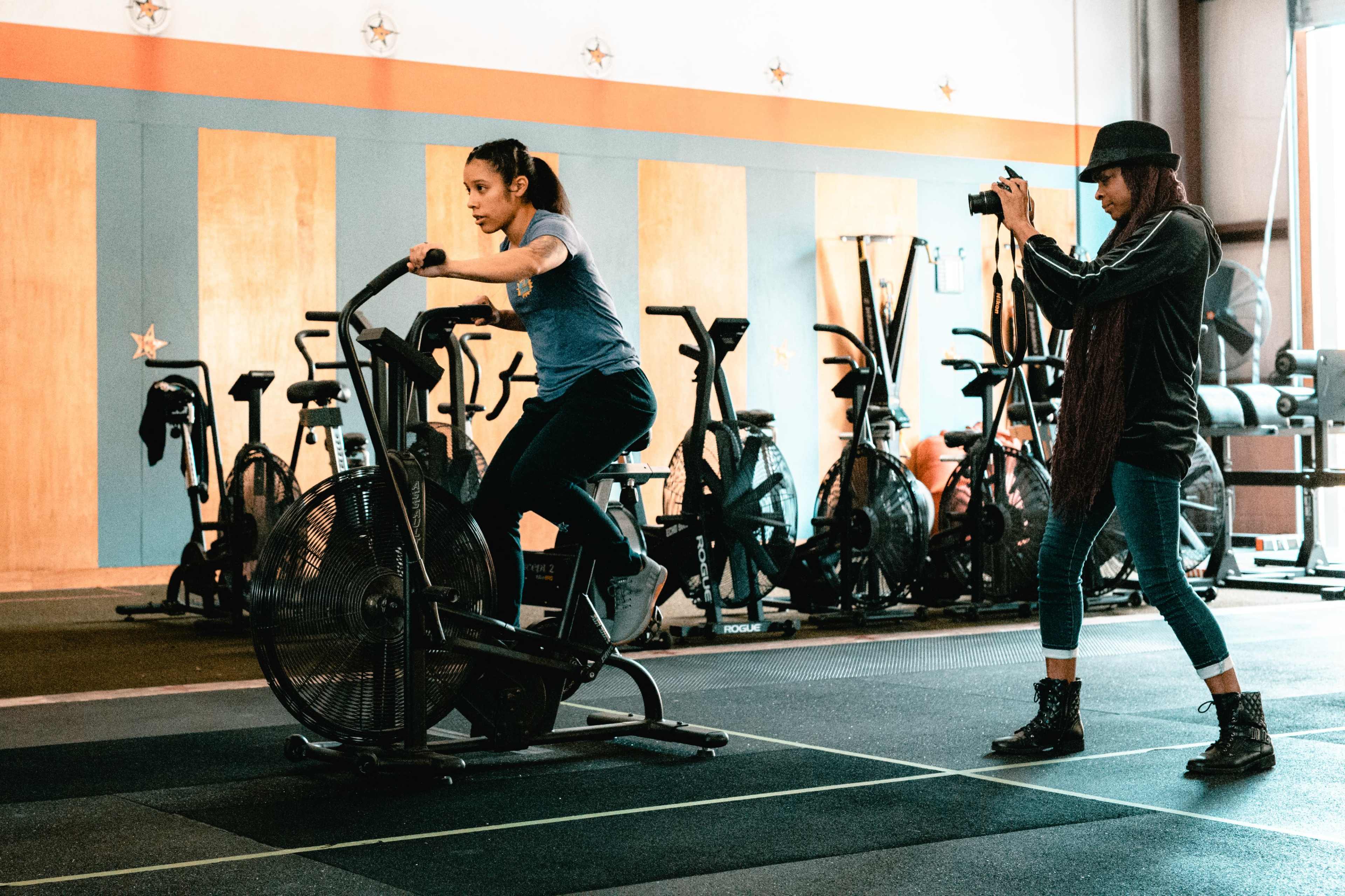 A woman exercises on a stationary bike while another woman photographs her in a gym setting.