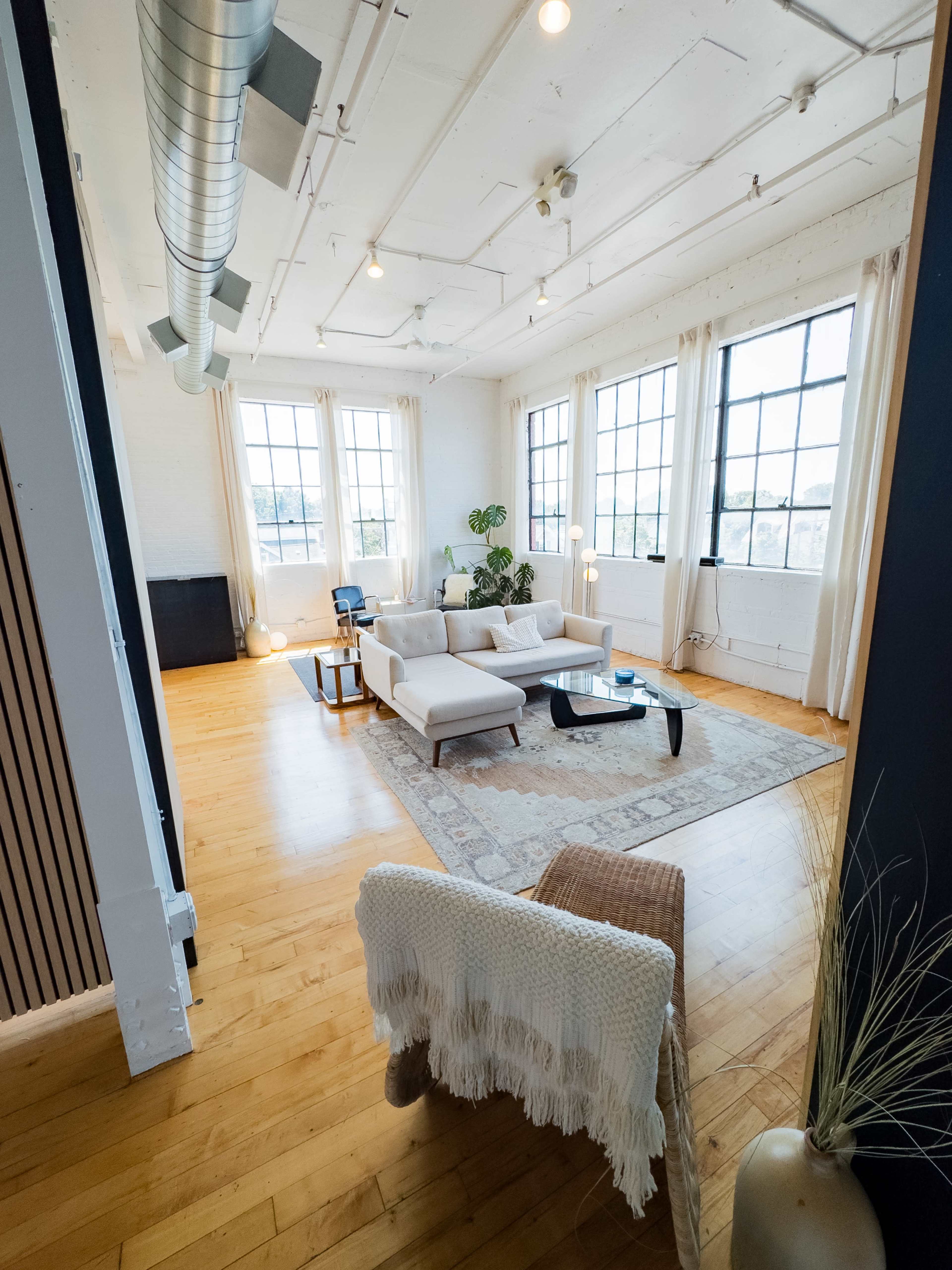 The image shows a spacious, well-lit living area featuring a modern sofa set, a coffee table, and large windows with natural light streaming in.