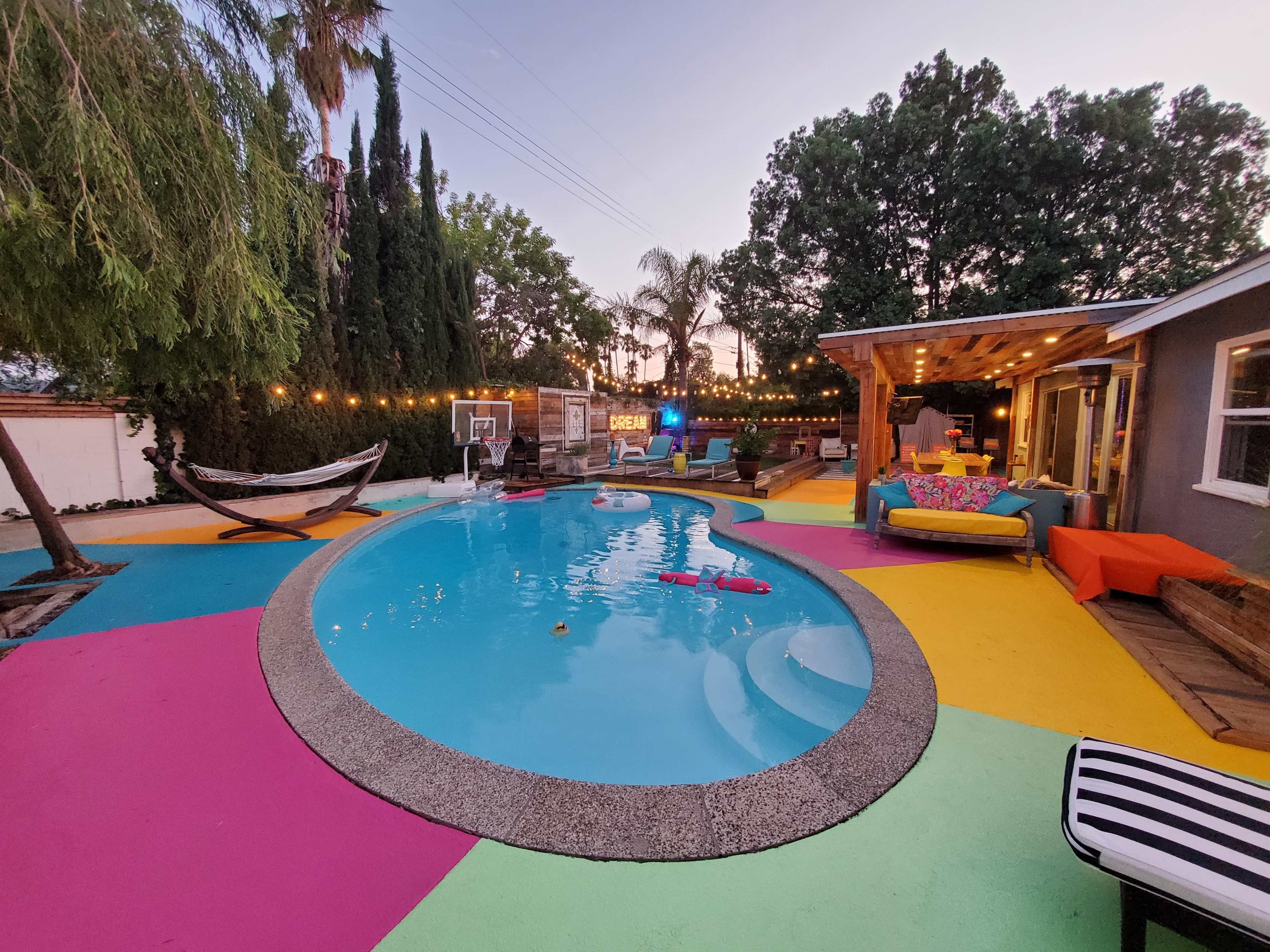 The scene features a vibrant backyard with a swimming pool, colorful deck, a hammock, and outdoor seating illuminated by string lights.