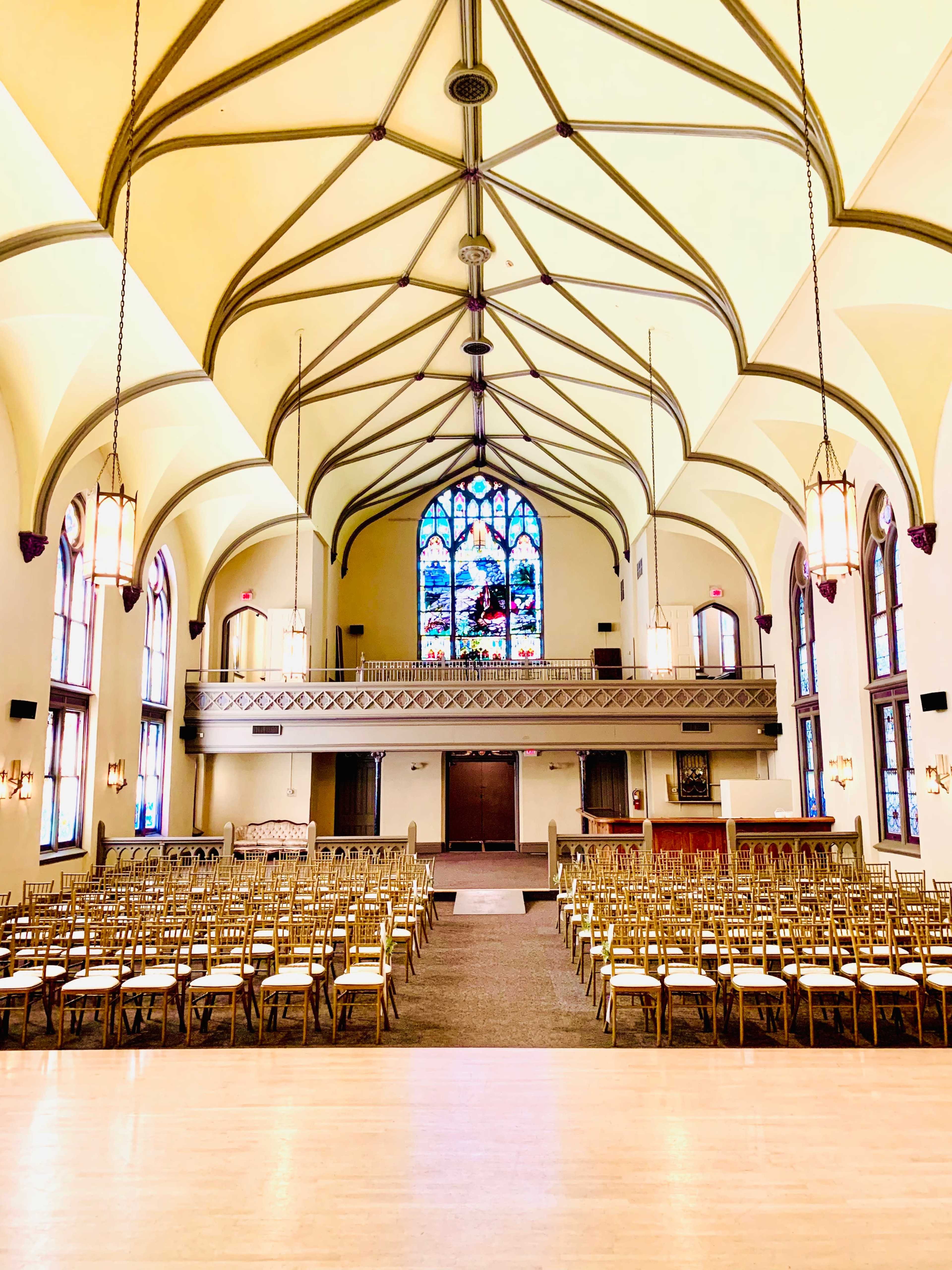 The interior of a church featuring a vaulted ceiling, stained glass windows, and rows of empty wooden chairs arranged for a gathering.