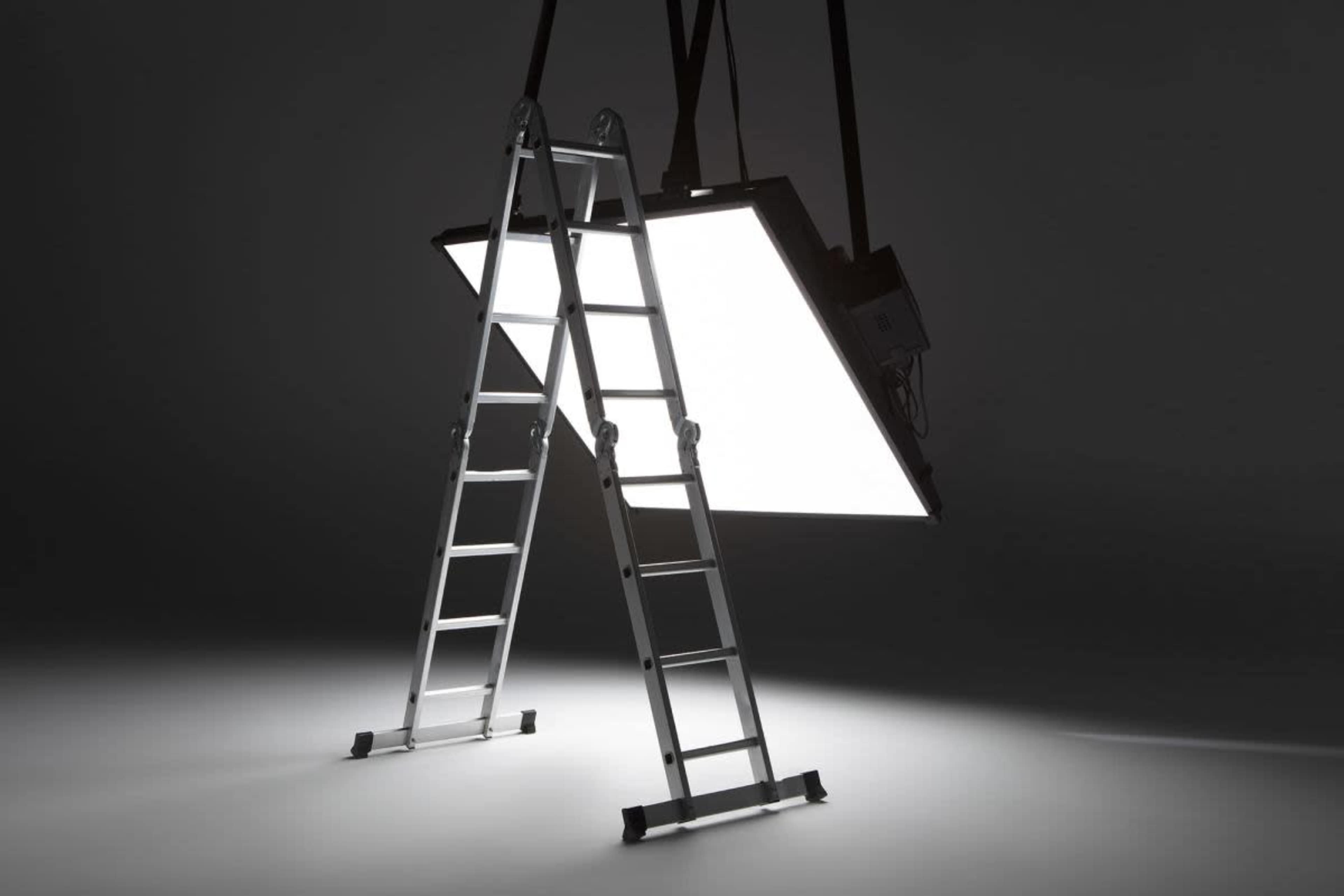 A metal ladder stands next to a large, bright rectangular light panel suspended from above in a dark space.