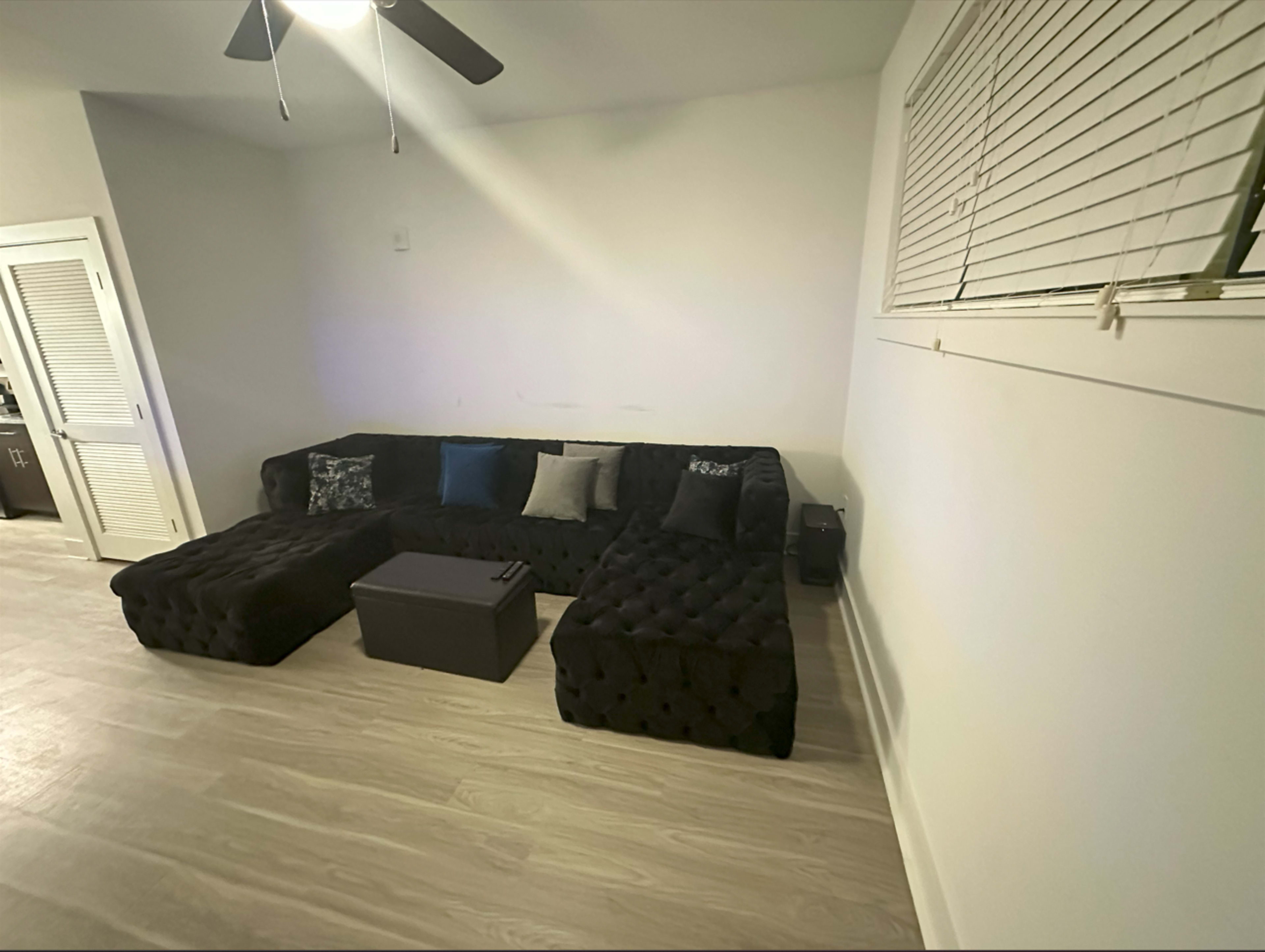 The image shows a black tufted sectional sofa with decorative pillows, positioned against a white wall in a room with light wood flooring.