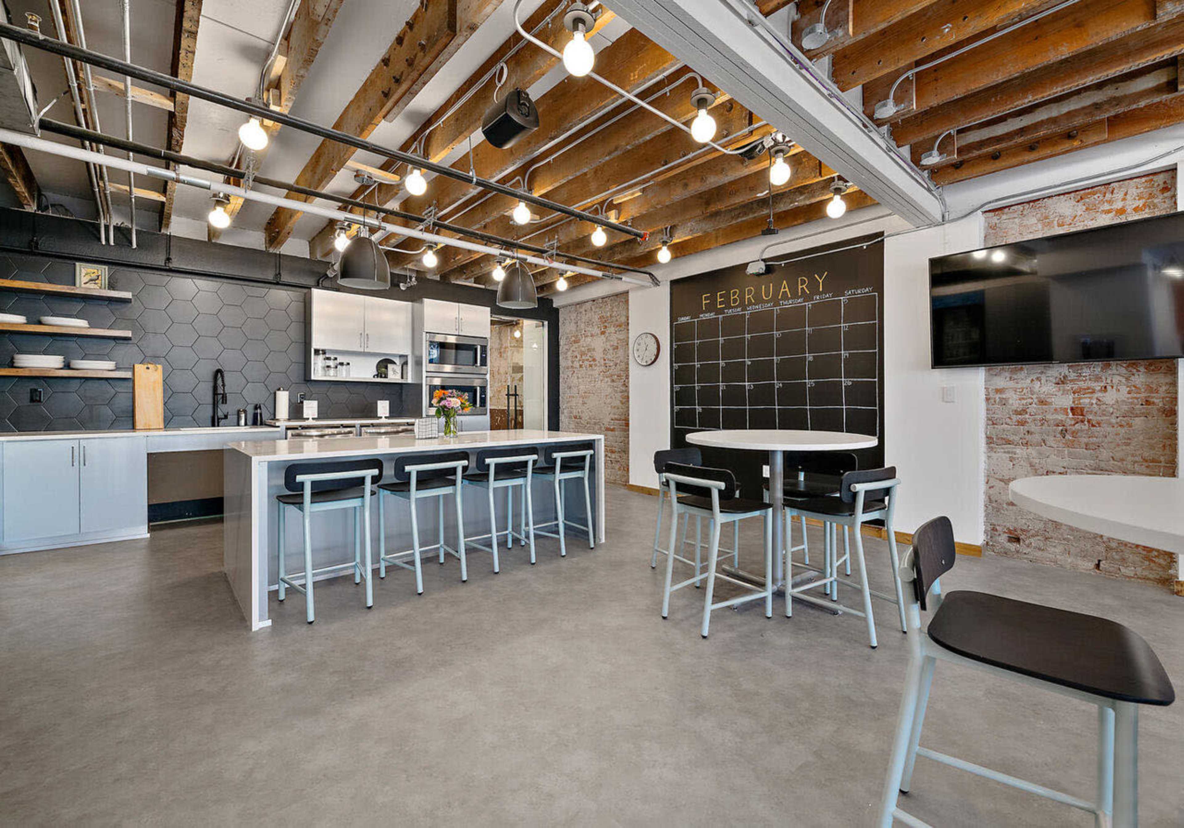 A modern kitchen and dining area with exposed beams, a large island, bar stools, and a chalkboard wall displaying the month of February.