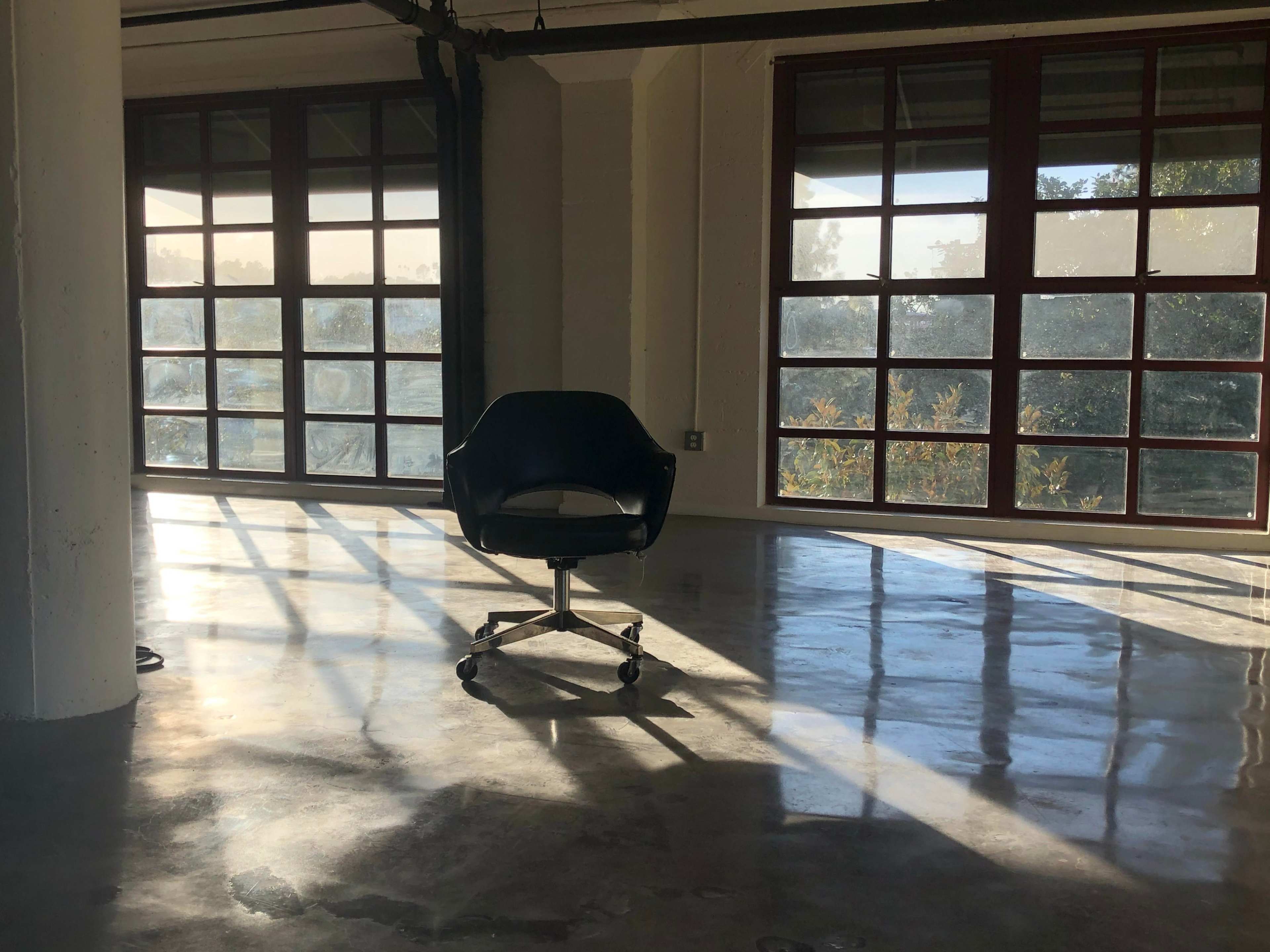 An empty office chair is positioned on a polished concrete floor, illuminated by sunlight streaming through large windows.