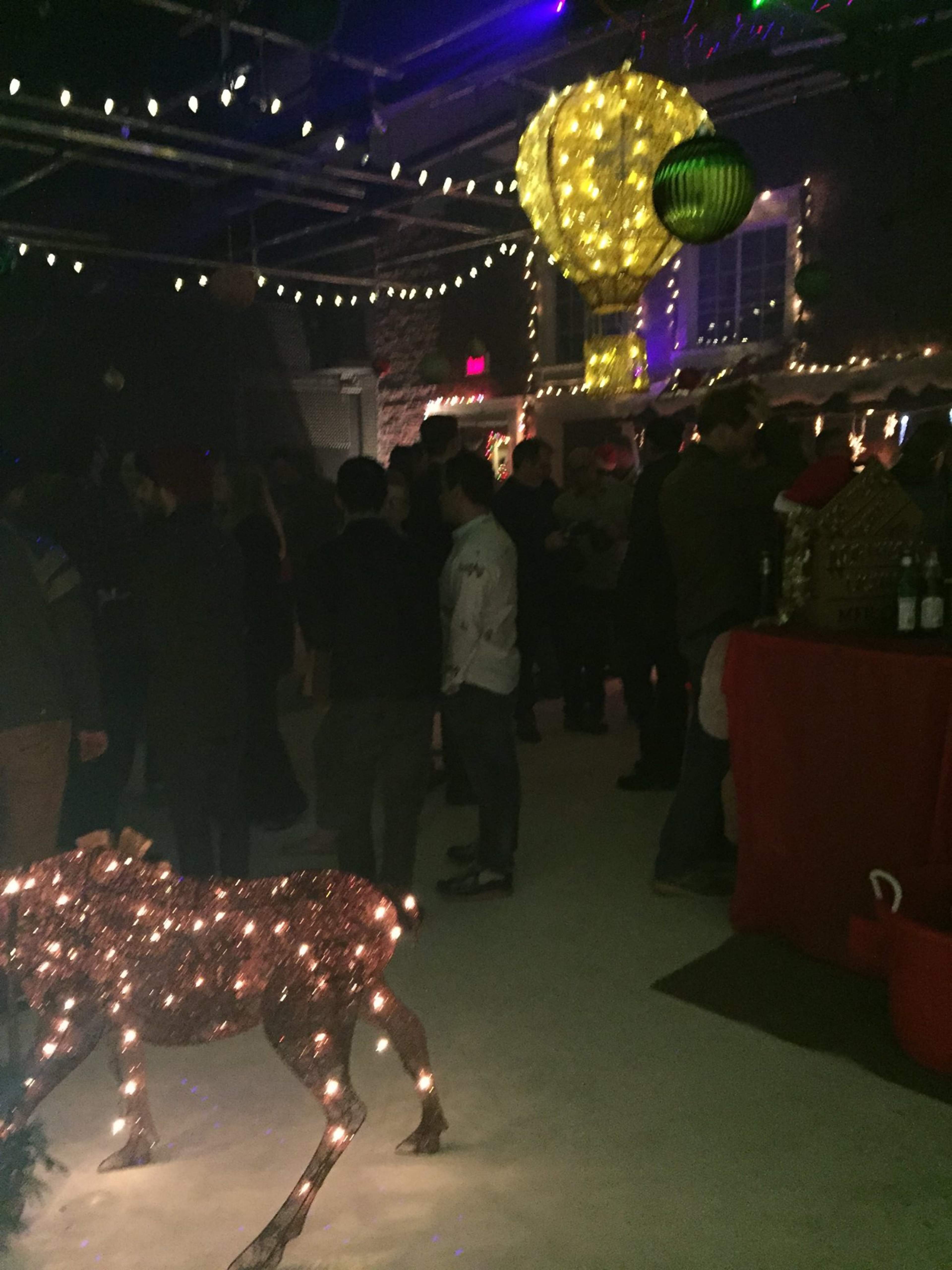 A crowded indoor venue features festive decorations, including a illuminated reindeer and a large hot air balloon, with guests mingling under string lights.