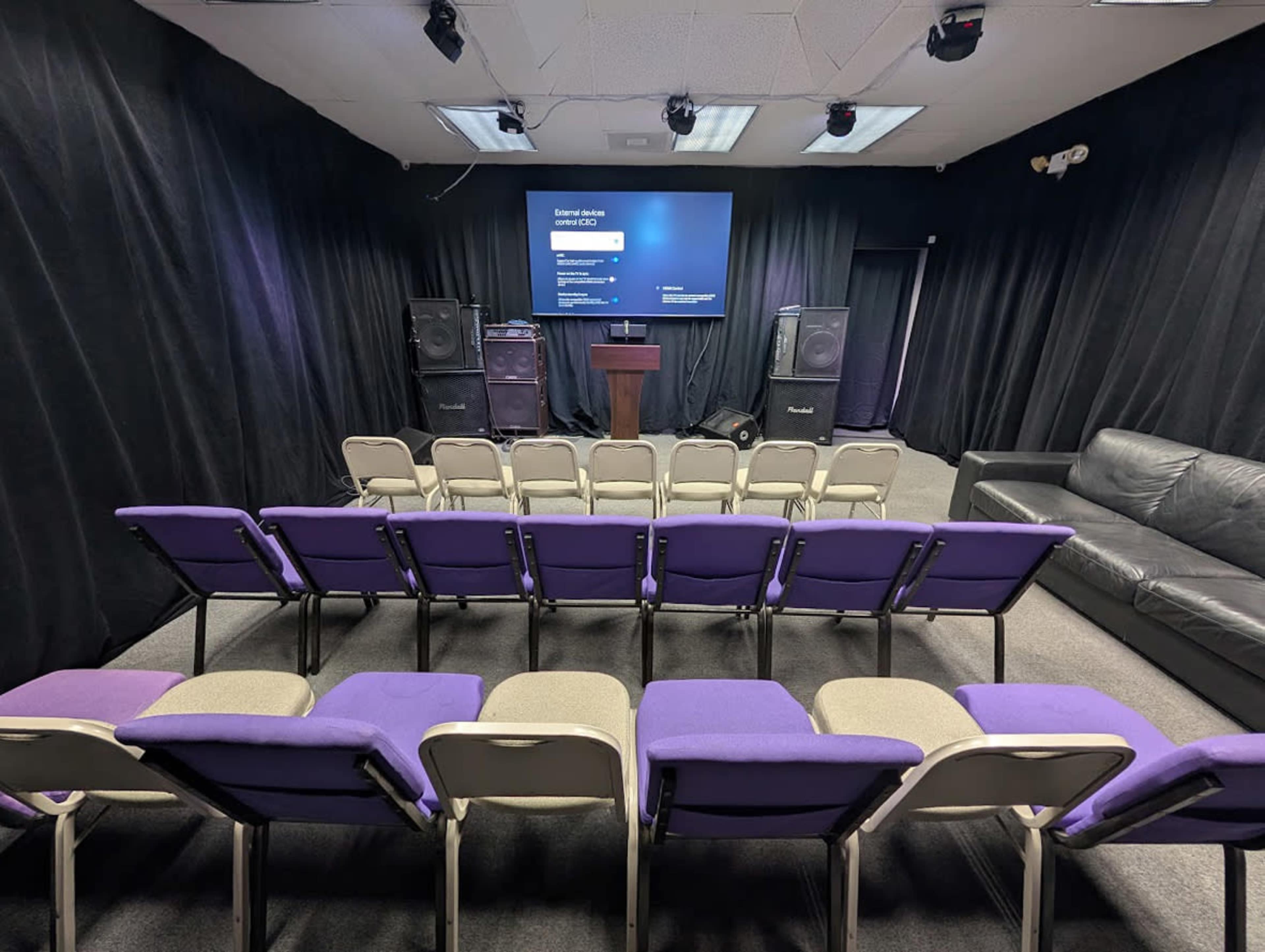 The image shows a seating area with purple and beige chairs arranged in rows facing a screen, flanked by large speakers and a podium in a darkened room.