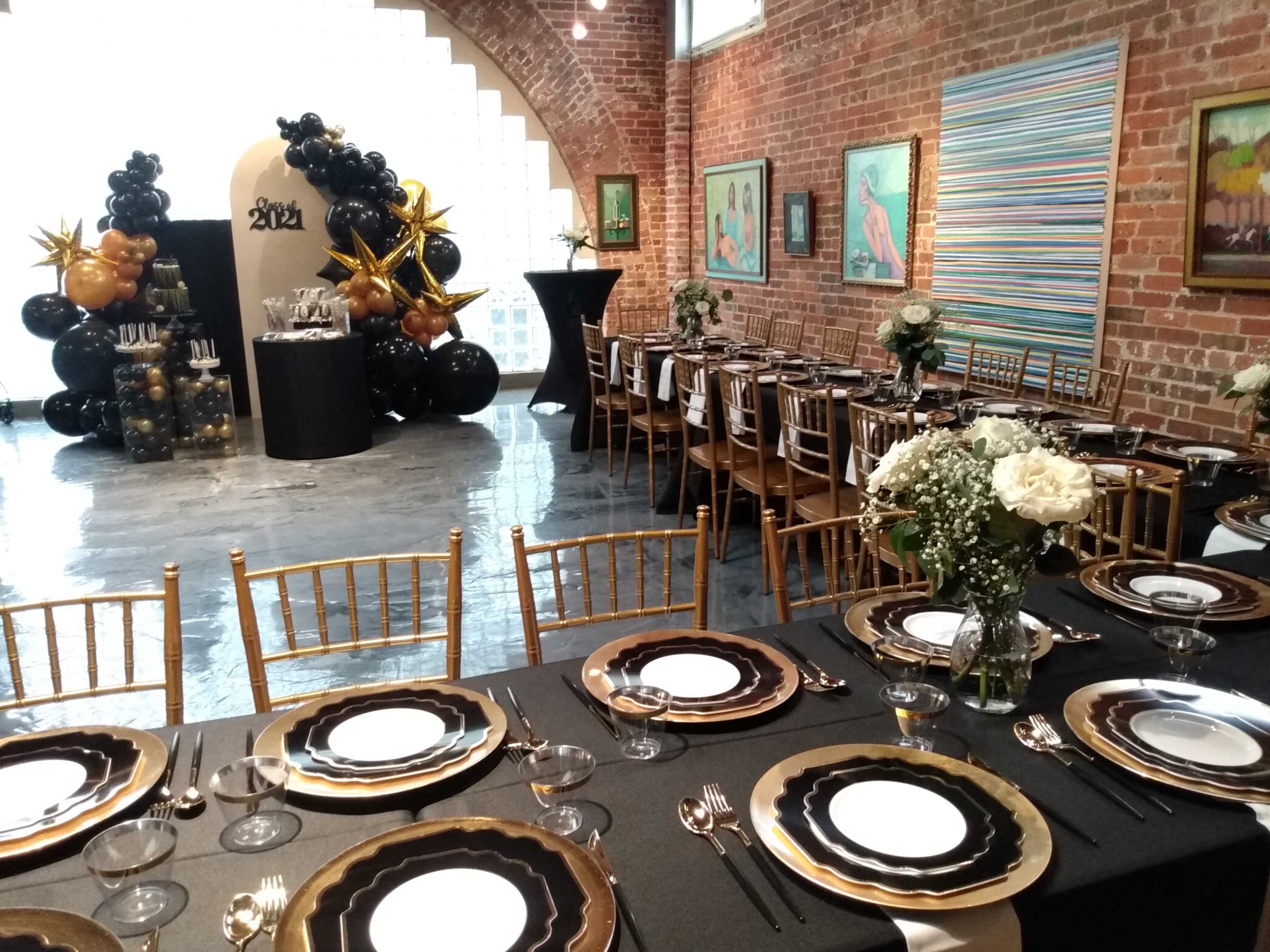 A decorated event space featuring a dining setup with black and gold table settings, surrounded by a backdrop of balloons and art on the walls.