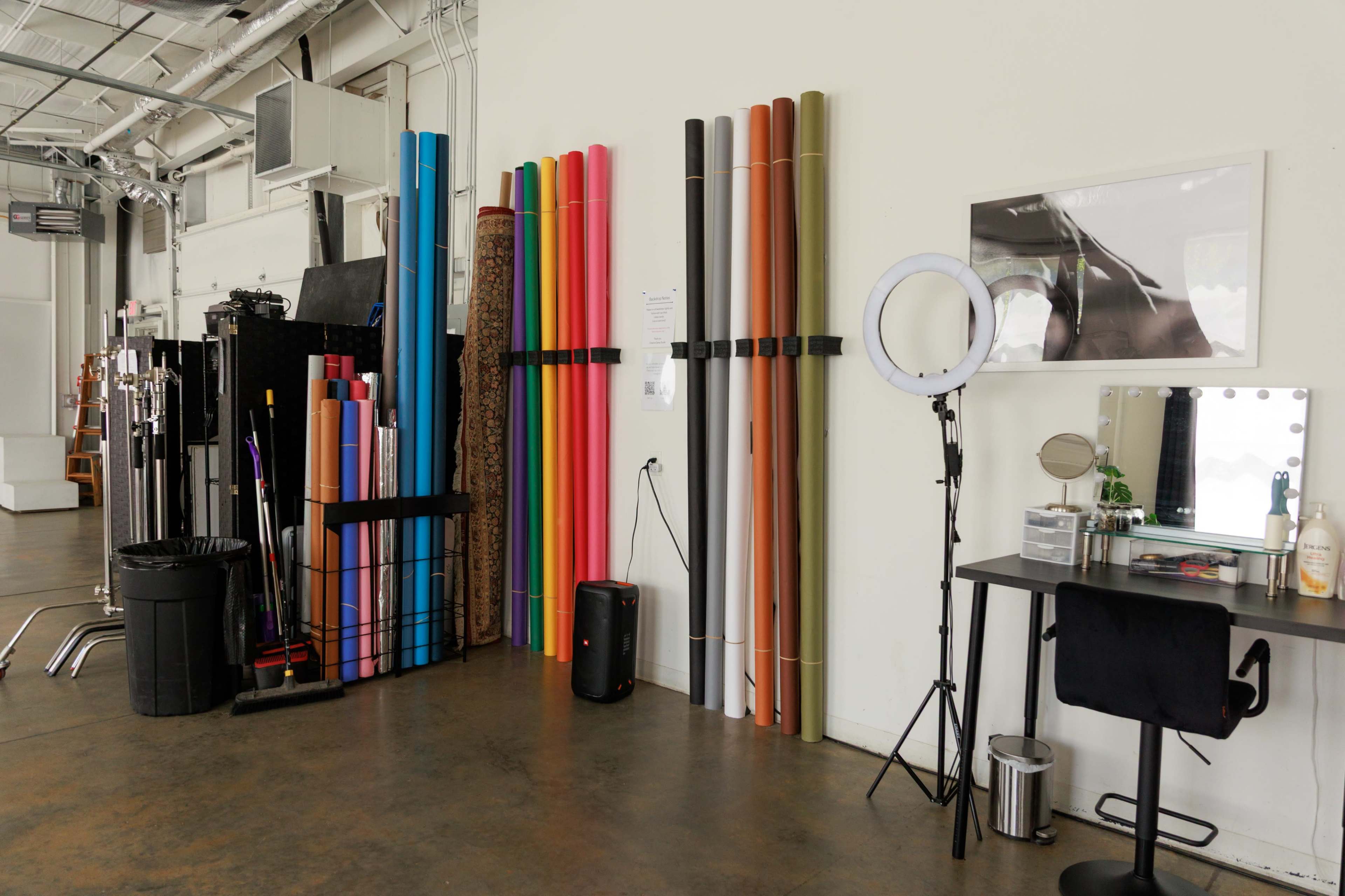 The image shows a studio space with colorful rolls of material stacked against the wall, a mirror and vanity setup, a ring light, and a trash bin nearby.
