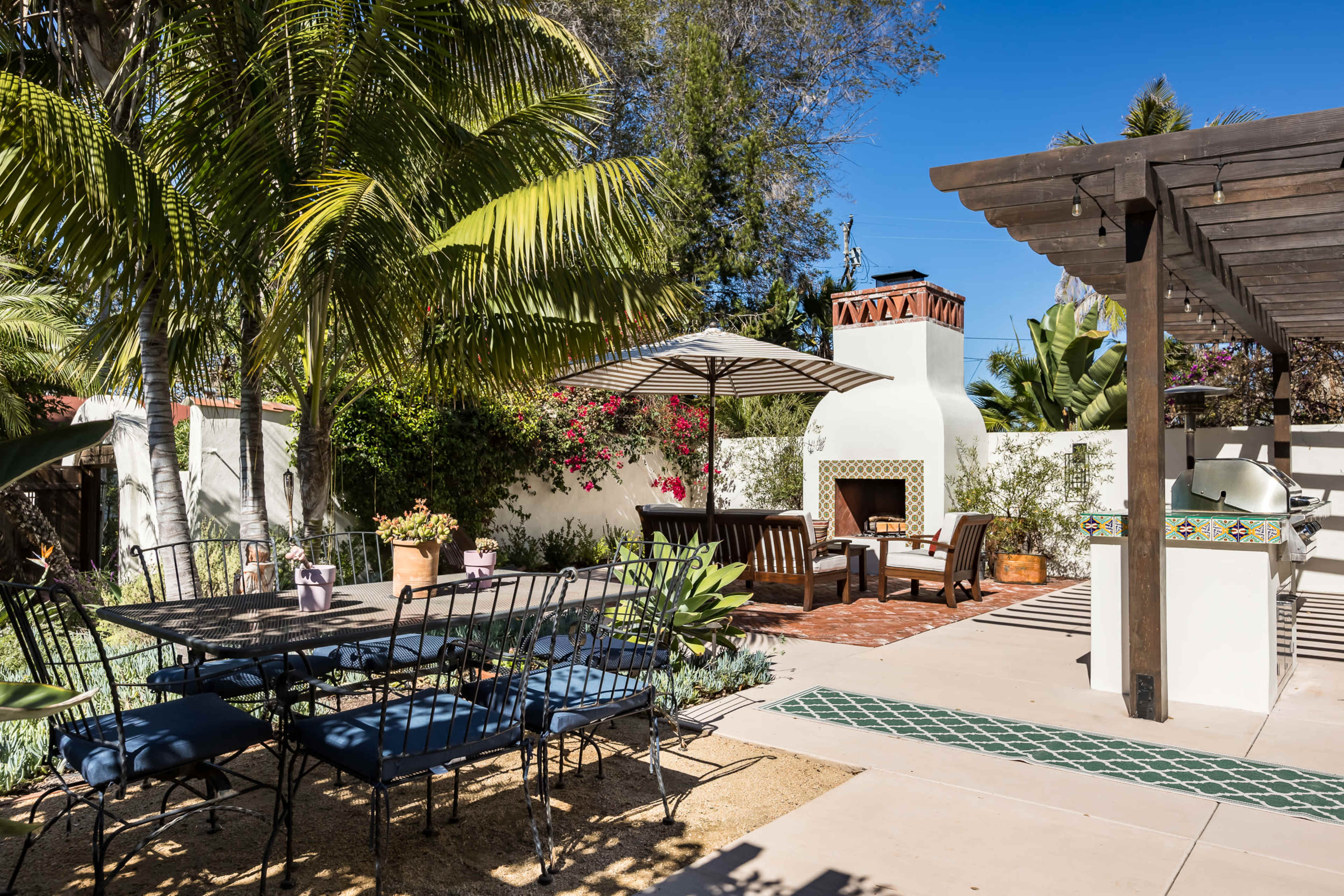 The image shows a landscaped outdoor patio area with a dining table, chairs, an umbrella, and a fireplace surrounded by lush greenery.