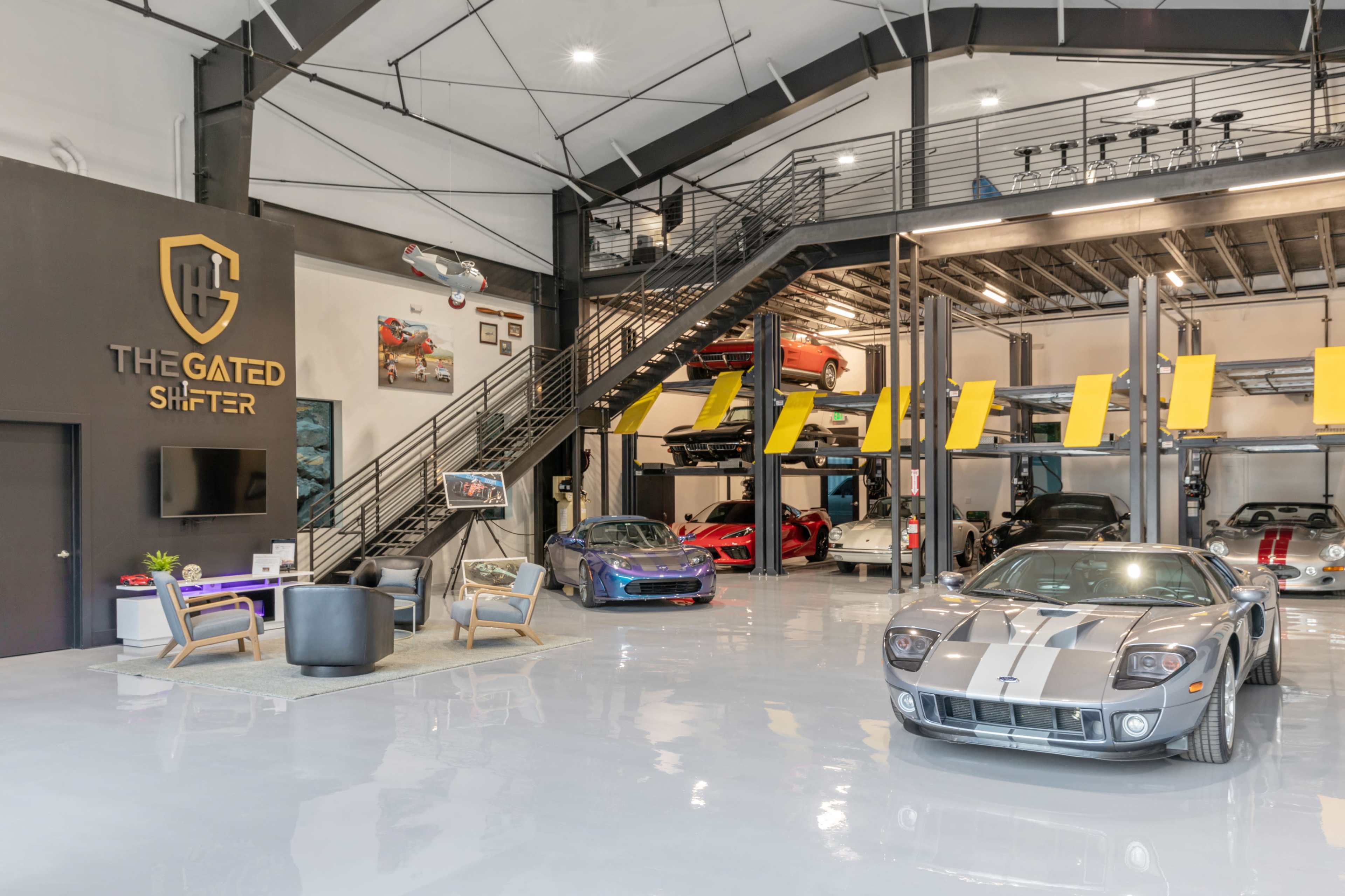 The image shows a modern car showroom with several luxury vehicles displayed on raised platforms, featuring a lounge area with seating and a large wall sign that reads "The Gated Shifter."