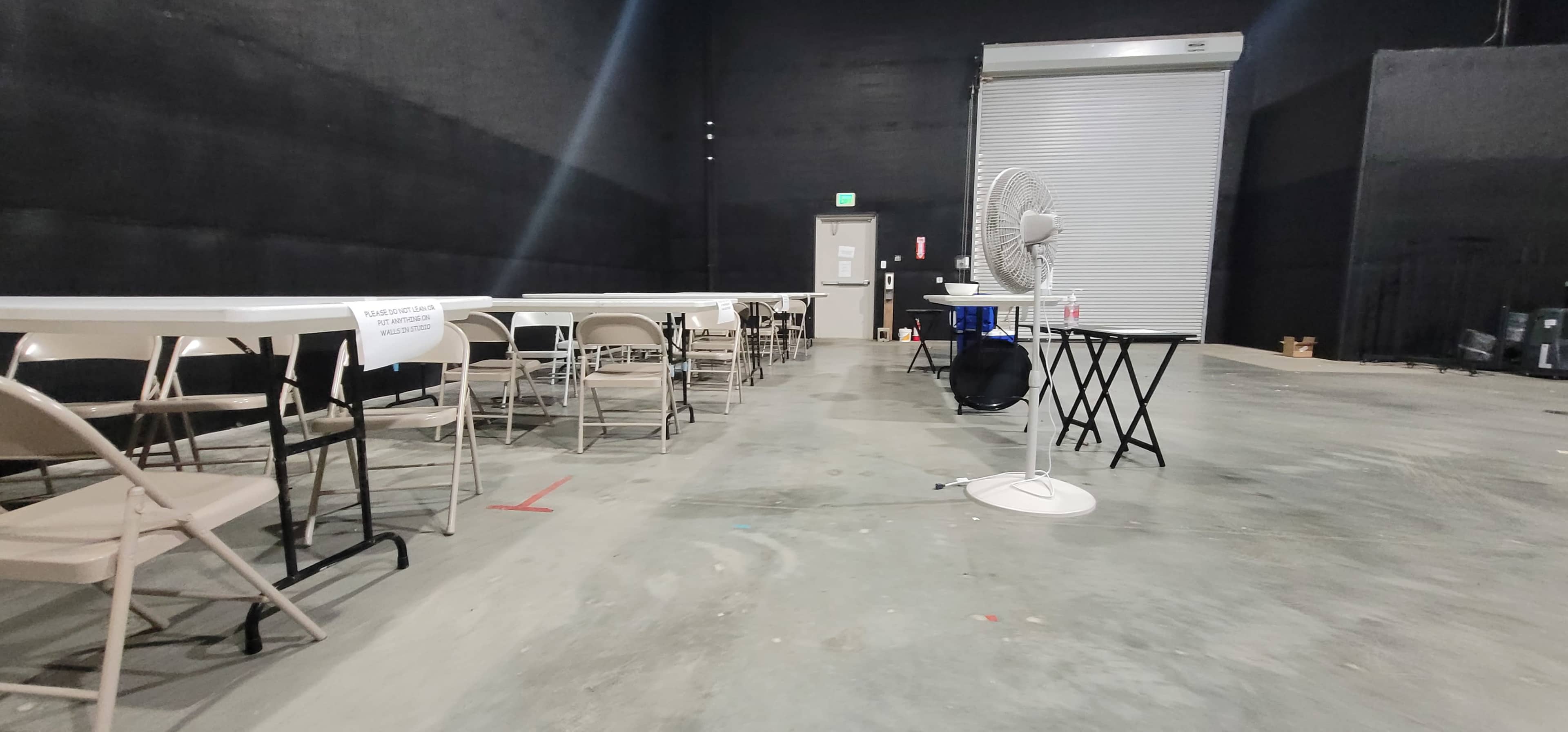 A spacious room with several tables and chairs arranged, a fan, and a closed rolling door in the background.