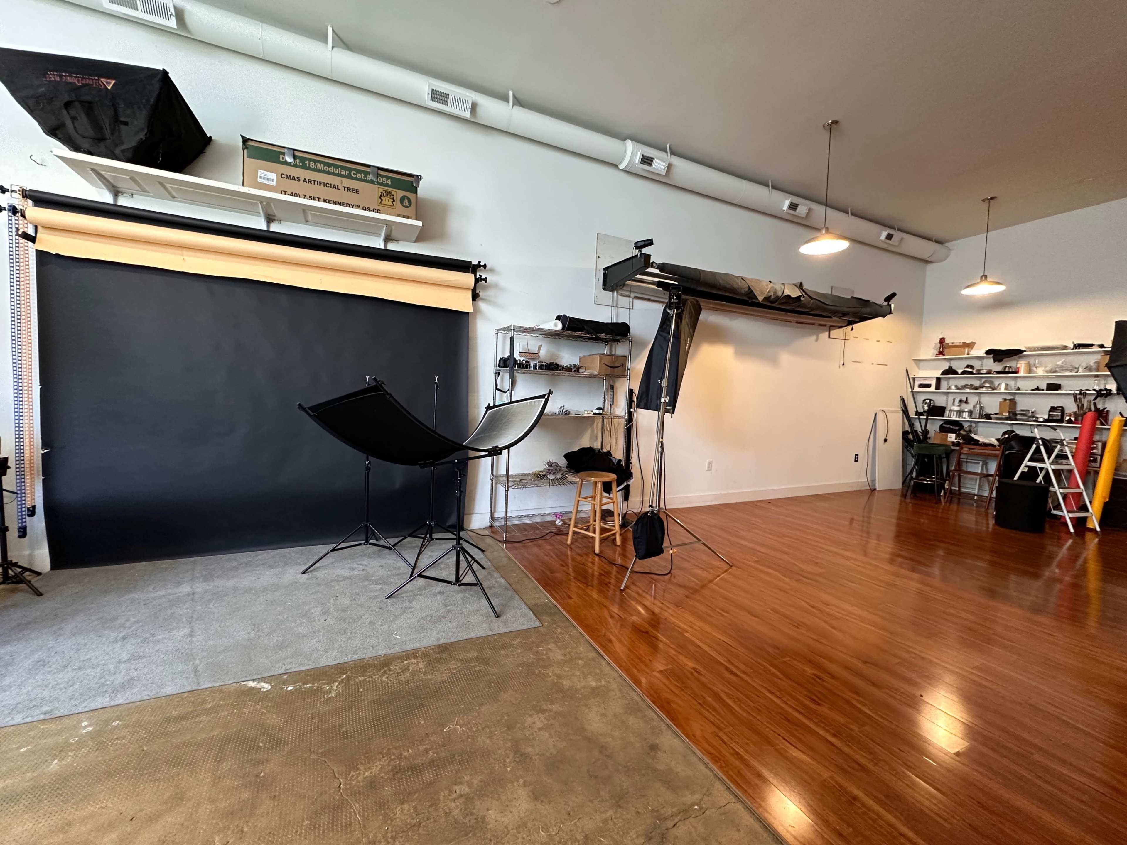 The image shows a photography studio with a black backdrop, lighting equipment, and various tools arranged on shelves.