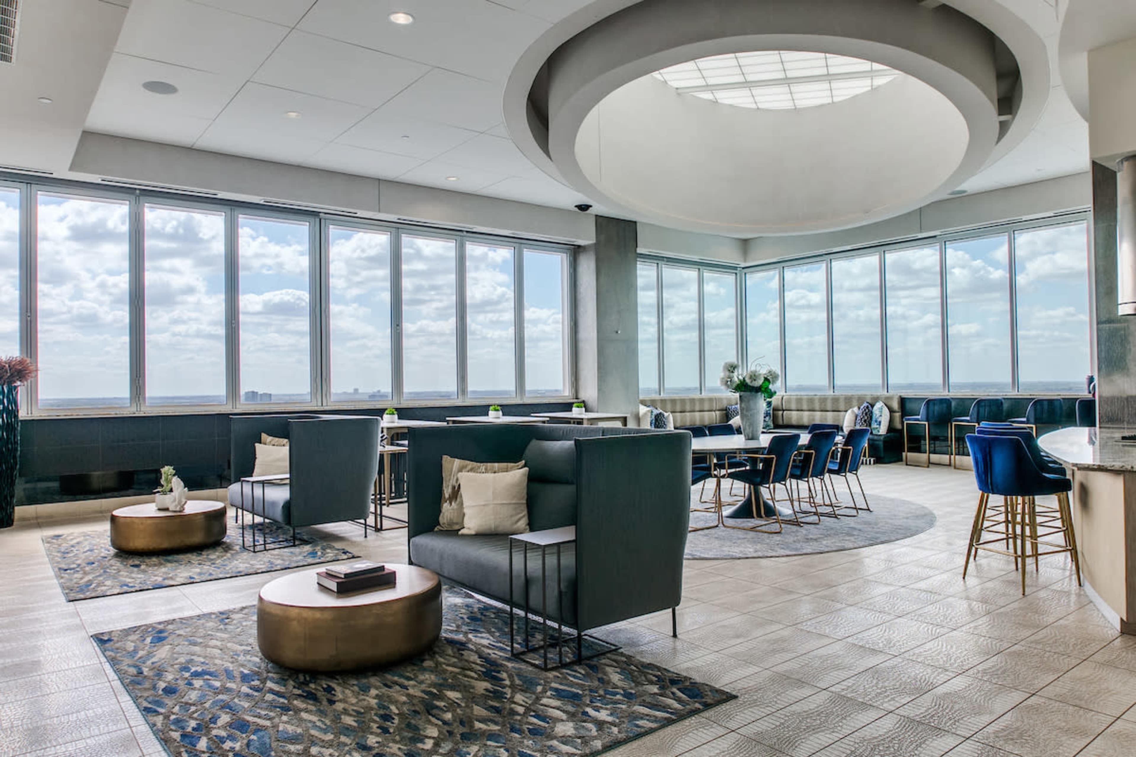 A modern lounge area features large windows with a view, comfortable seating, and decorative rugs.