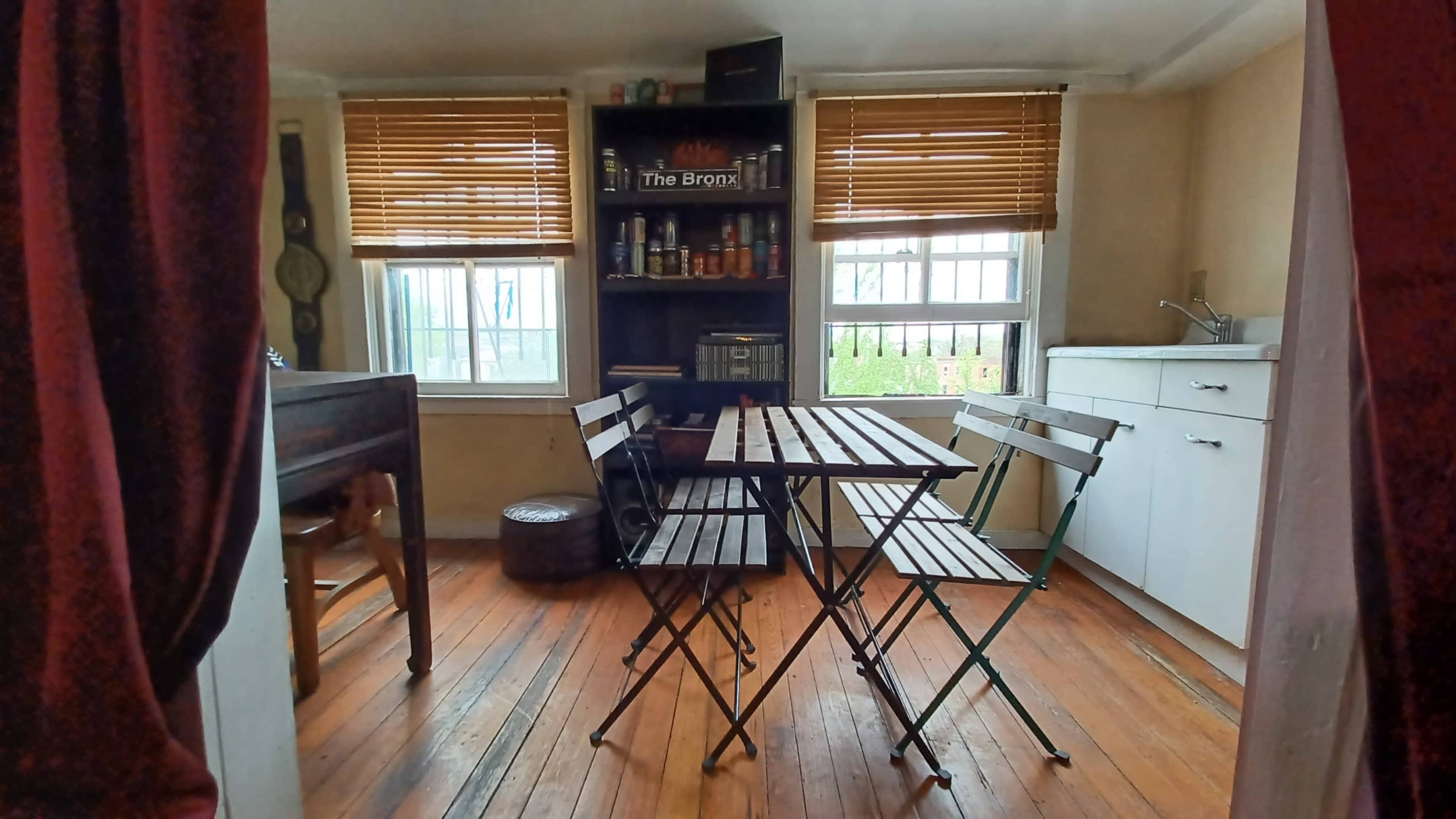 The image shows a small dining area with a wooden table and four folding chairs, surrounded by shelves and a kitchenette.