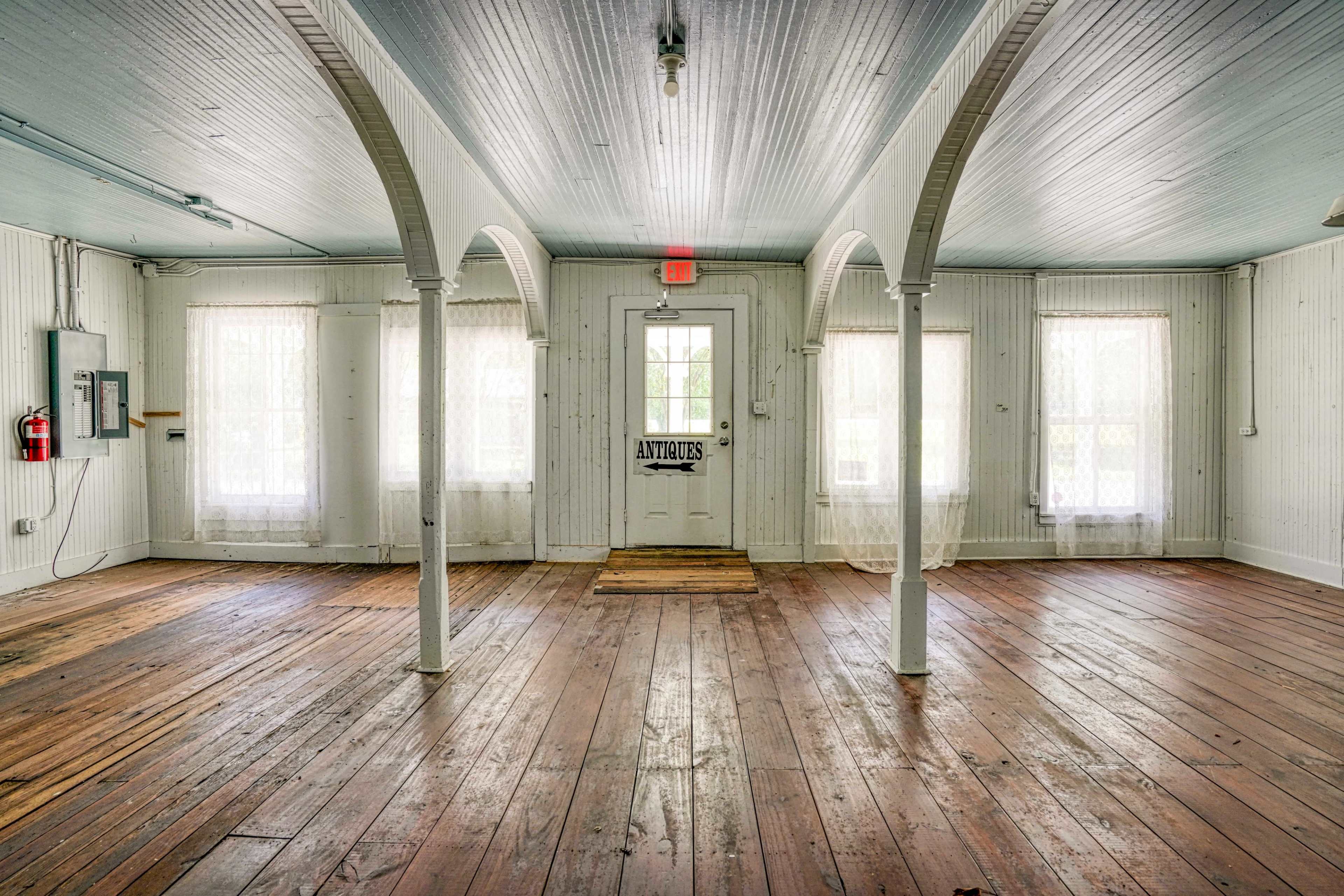 The image features an empty room with wooden floors, white walls, and large windows, highlighted by decorative arches leading to a door marked "Antiques."