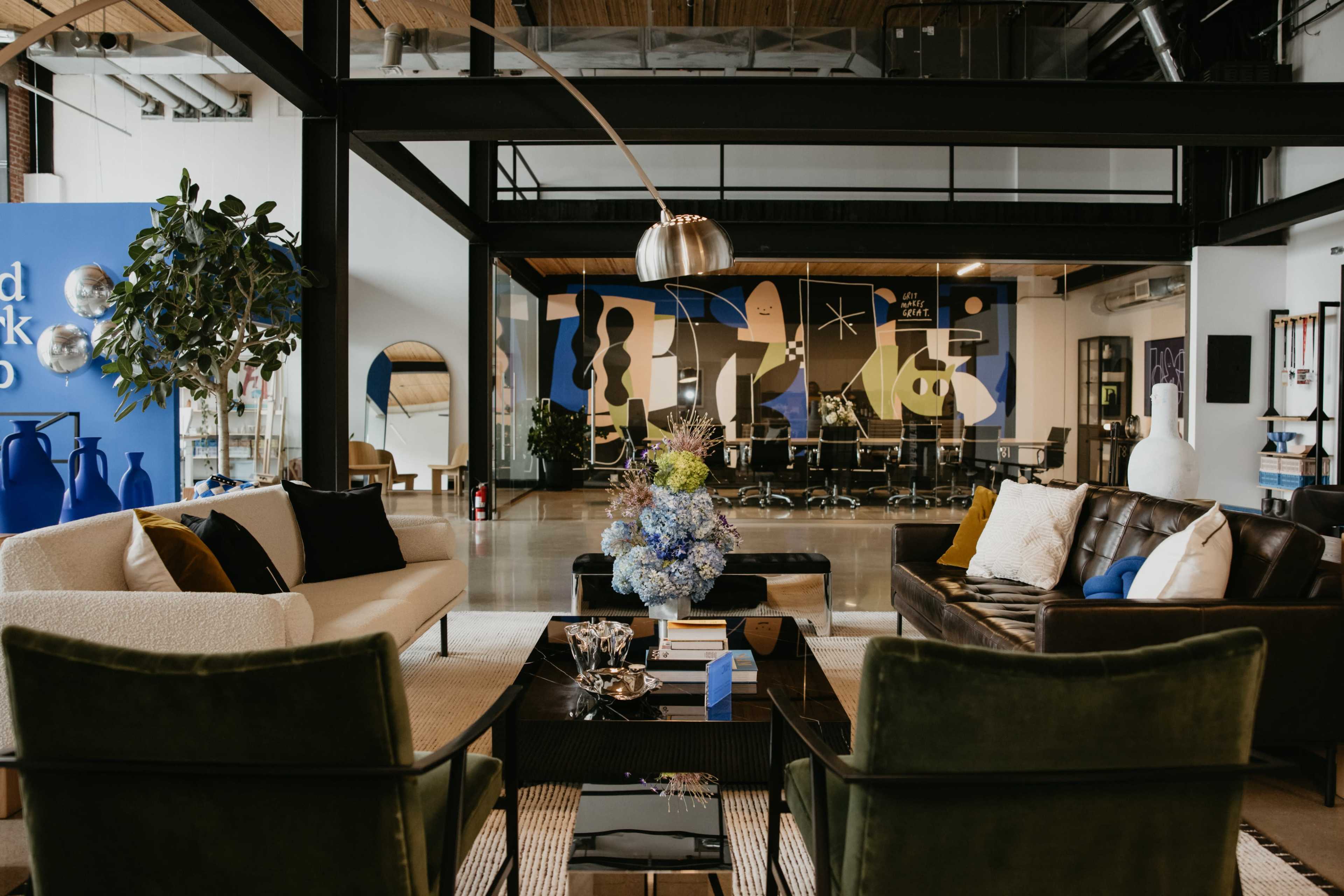 The image shows a modern office lounge area with two sofas, a coffee table adorned with flowers, and large colorful murals on the walls.