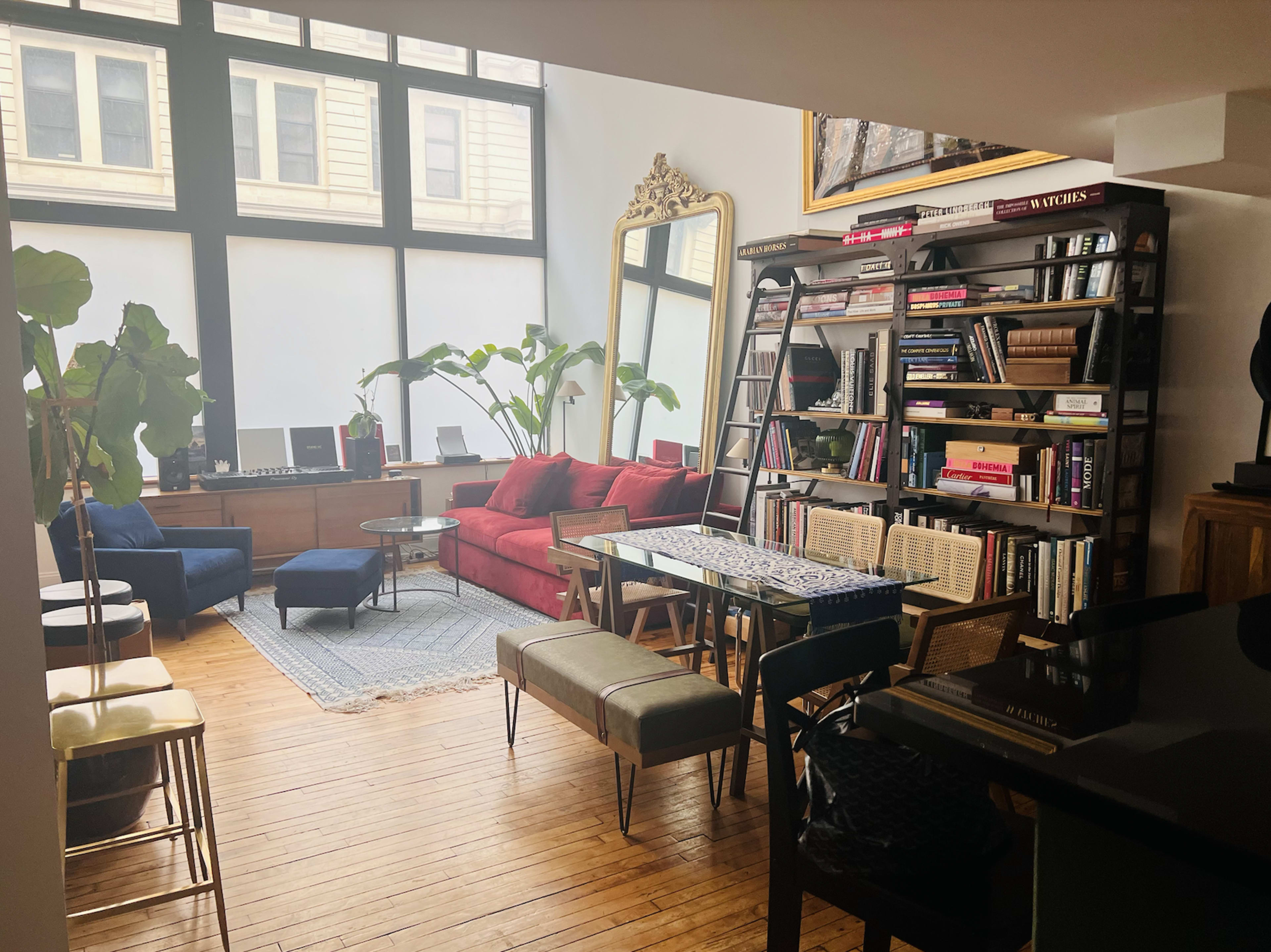 A contemporary living room with a mix of colorful furniture, large windows allowing natural light, and a bookshelf filled with books.