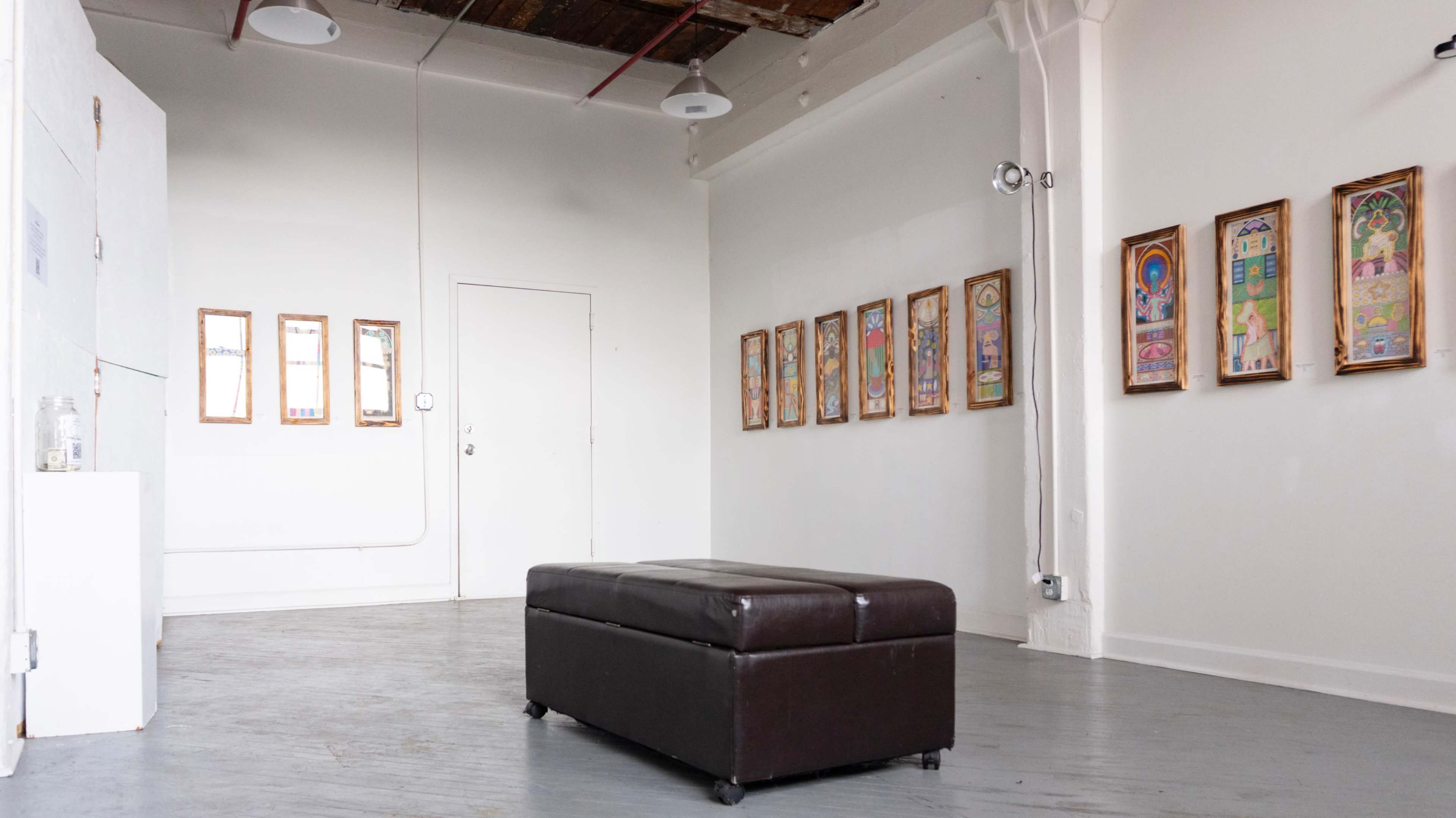 The image shows an art gallery with framed artworks hanging on the wall, a brown ottoman in the center, and a white door in the background.