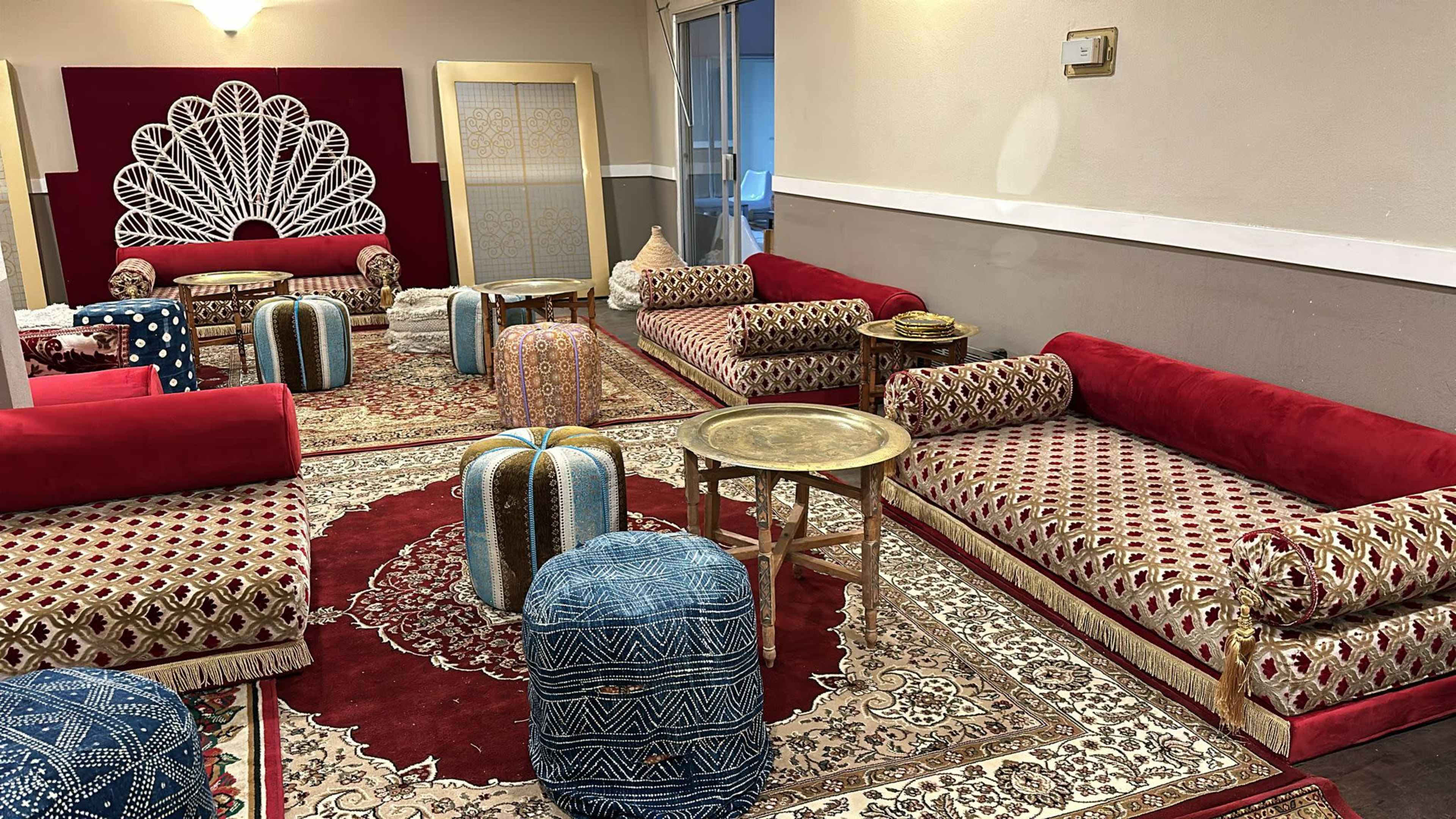 The image shows a seating area decorated with red patterned couches, colorful poufs, and a traditional low table on an ornate rug.