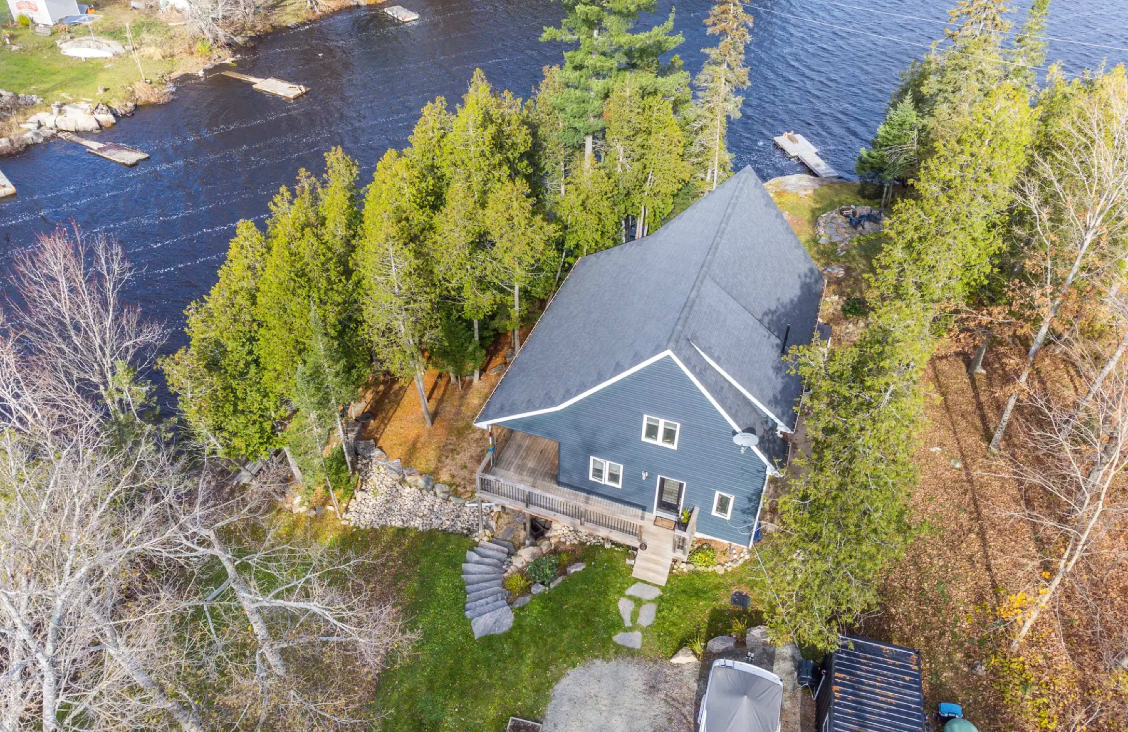 An aerial view of a blue house surrounded by trees near a river, with a landscaped yard and a gravel driveway.